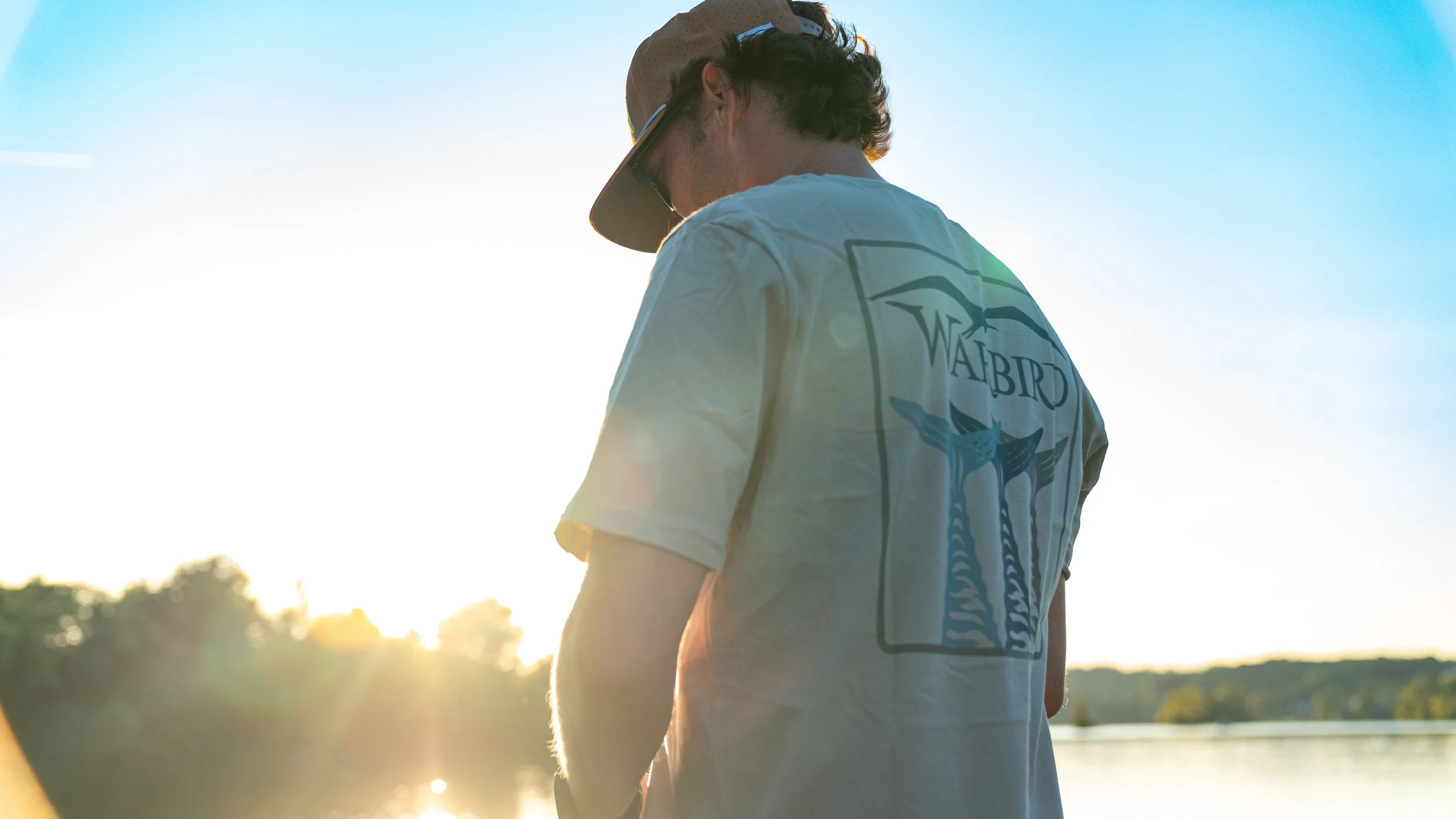 A person wearing a T-shirt with a bird logo and the word 'WABIRD' on the back, sunglasses, and a cap, standing outdoors near a body of water during sunset.