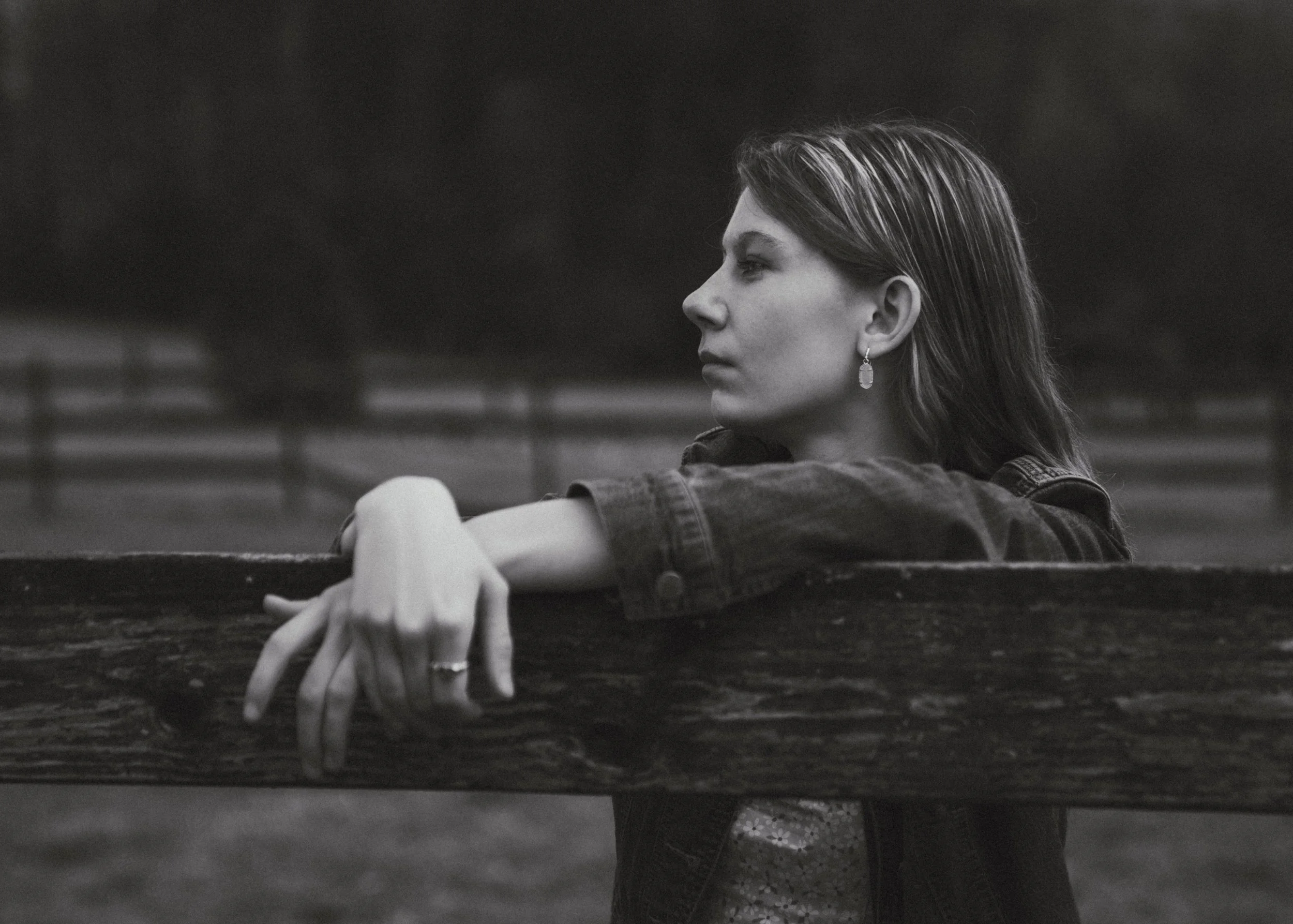 Black-and-white photograph of a woman with shoulder-length hair, wearing a denim jacket and earrings, resting her arms on a wooden fence, looking into the distance with a contemplative expression.