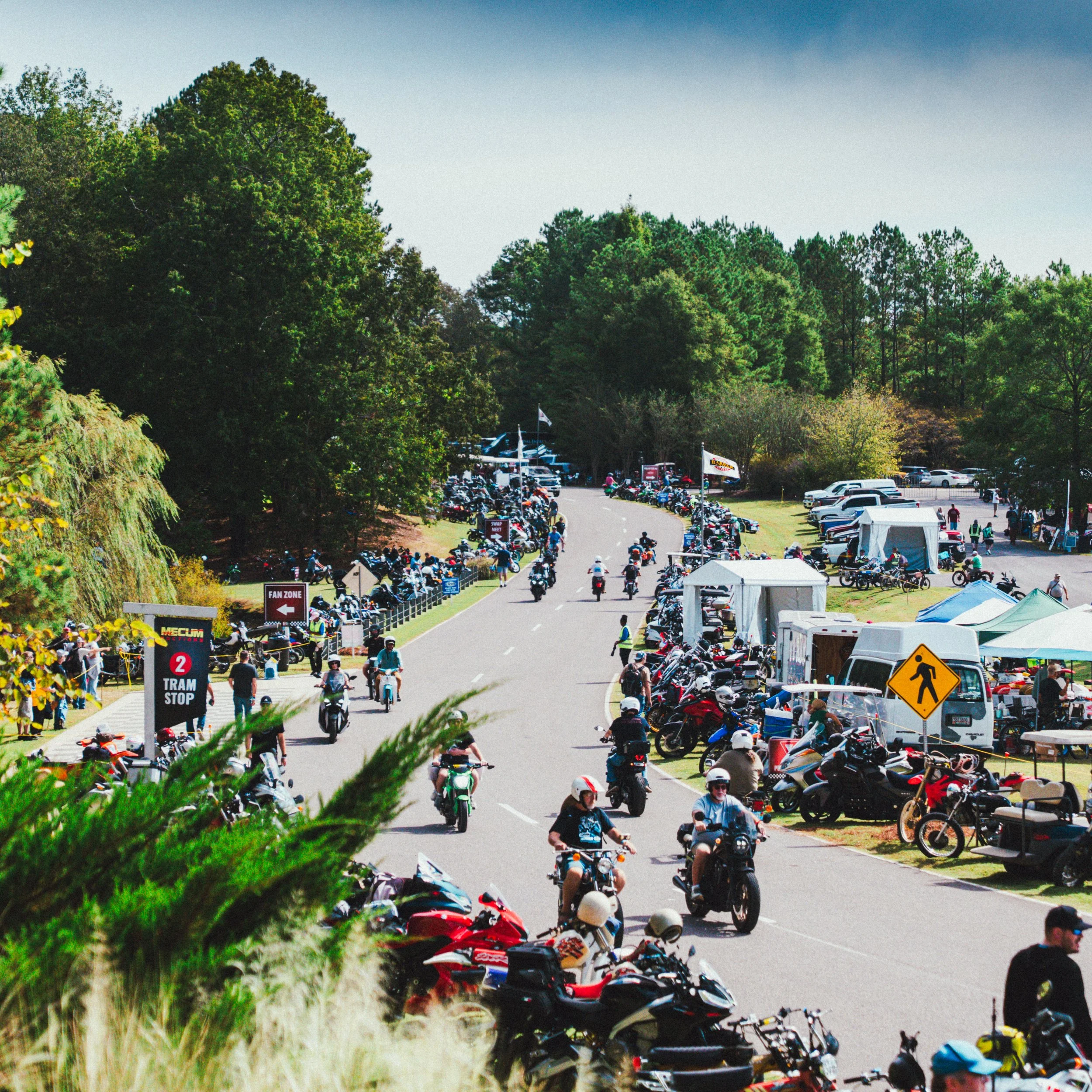 Crowd of motorcycles and people at a motorcycle event with tents and signs along a curved road, surrounded by green trees.