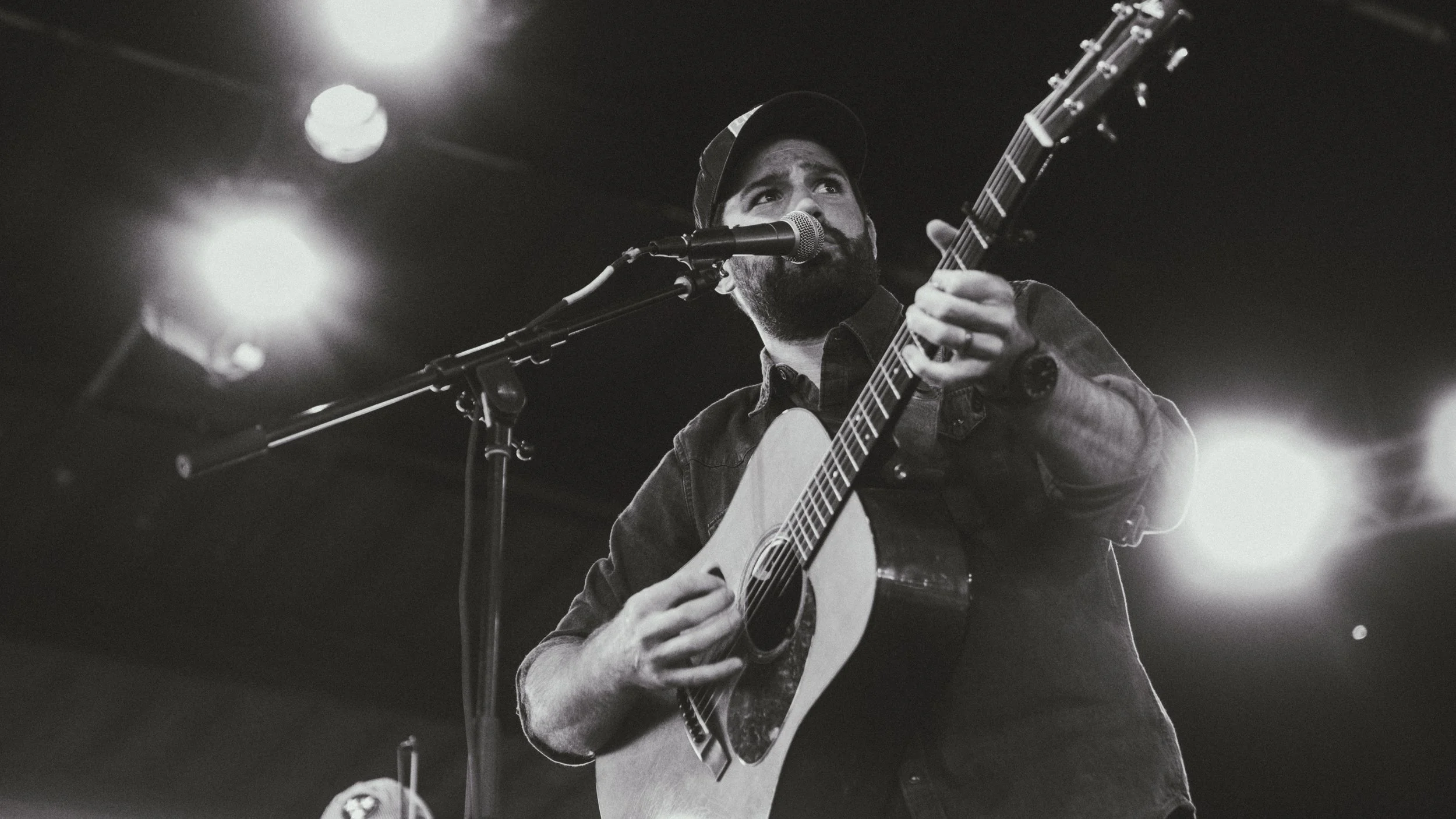 A man with a beard playing an acoustic guitar and singing into a microphone on stage, with bright lights in the background.