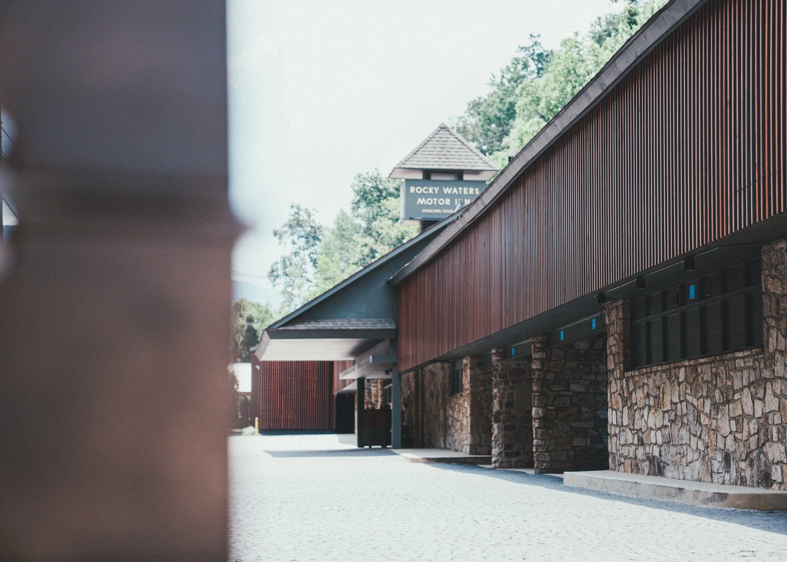 Side view of a building with stone and wooden exterior, with a sign indicating 'Rocky Waters Motor Inn' visible in the background, trees behind the building.