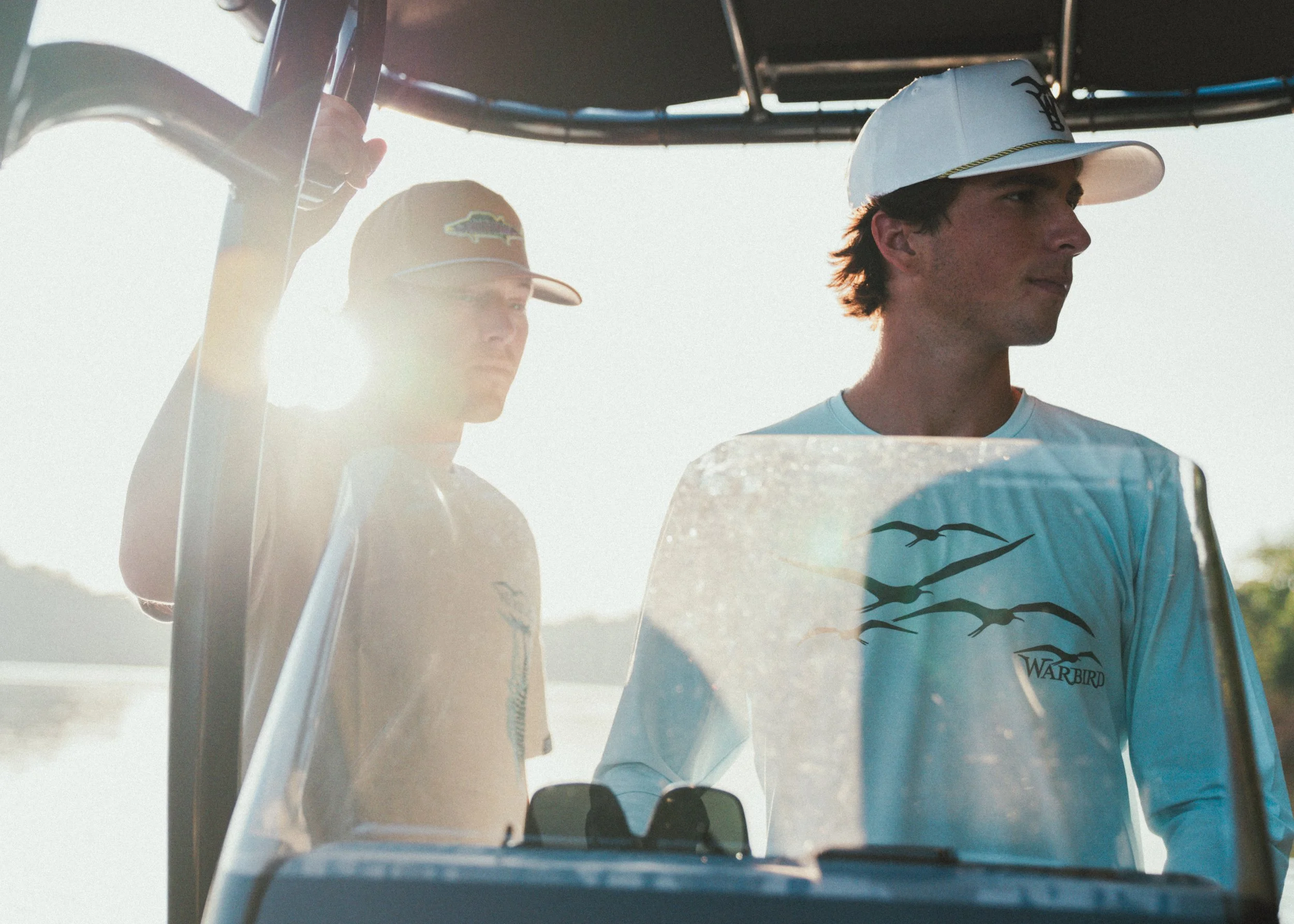 Two young men standing on a boat, wearing baseball caps, with one wearing sunglasses, in bright sunlight.