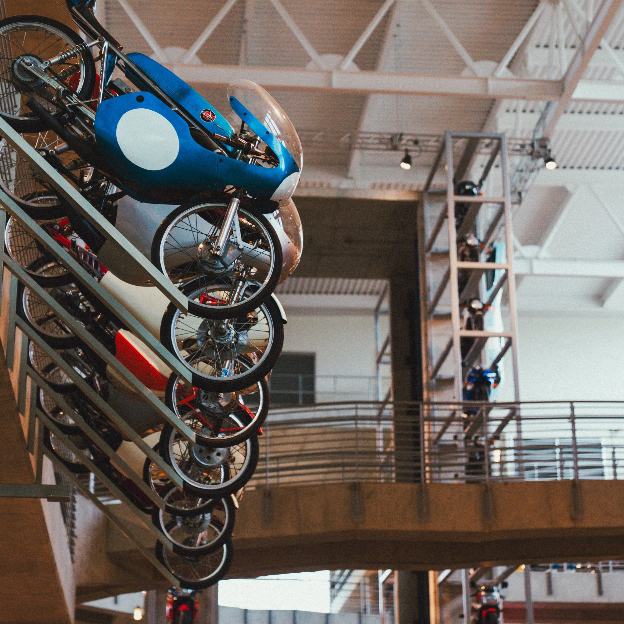 Multiple racing motorcycles mounted on a vertical display in a modern showroom or museum, with a wooden balcony and industrial ceiling in the background.
