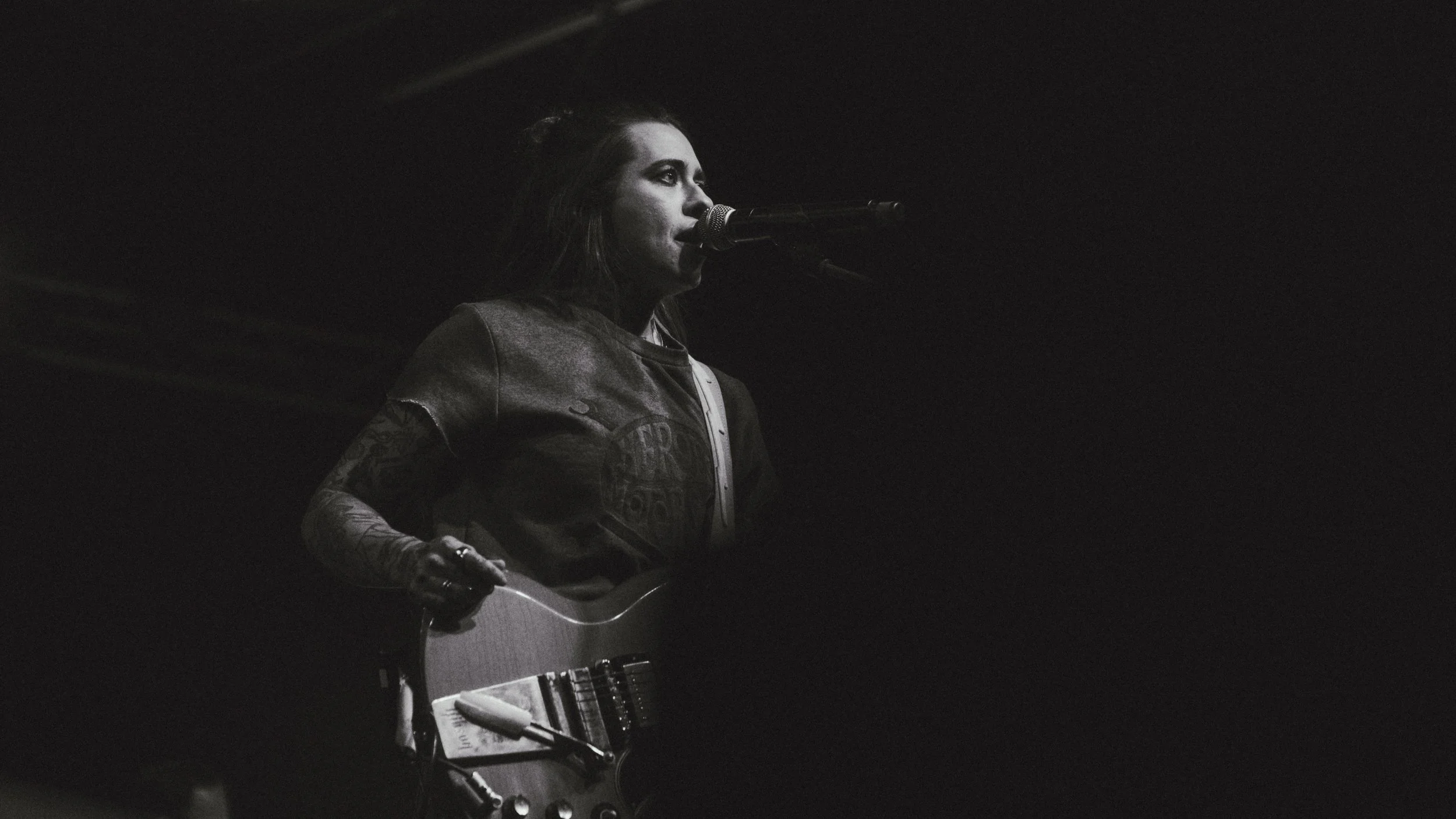 A female musician performing on stage, playing an electric guitar and singing into a microphone, in a dark setting with minimal lighting.