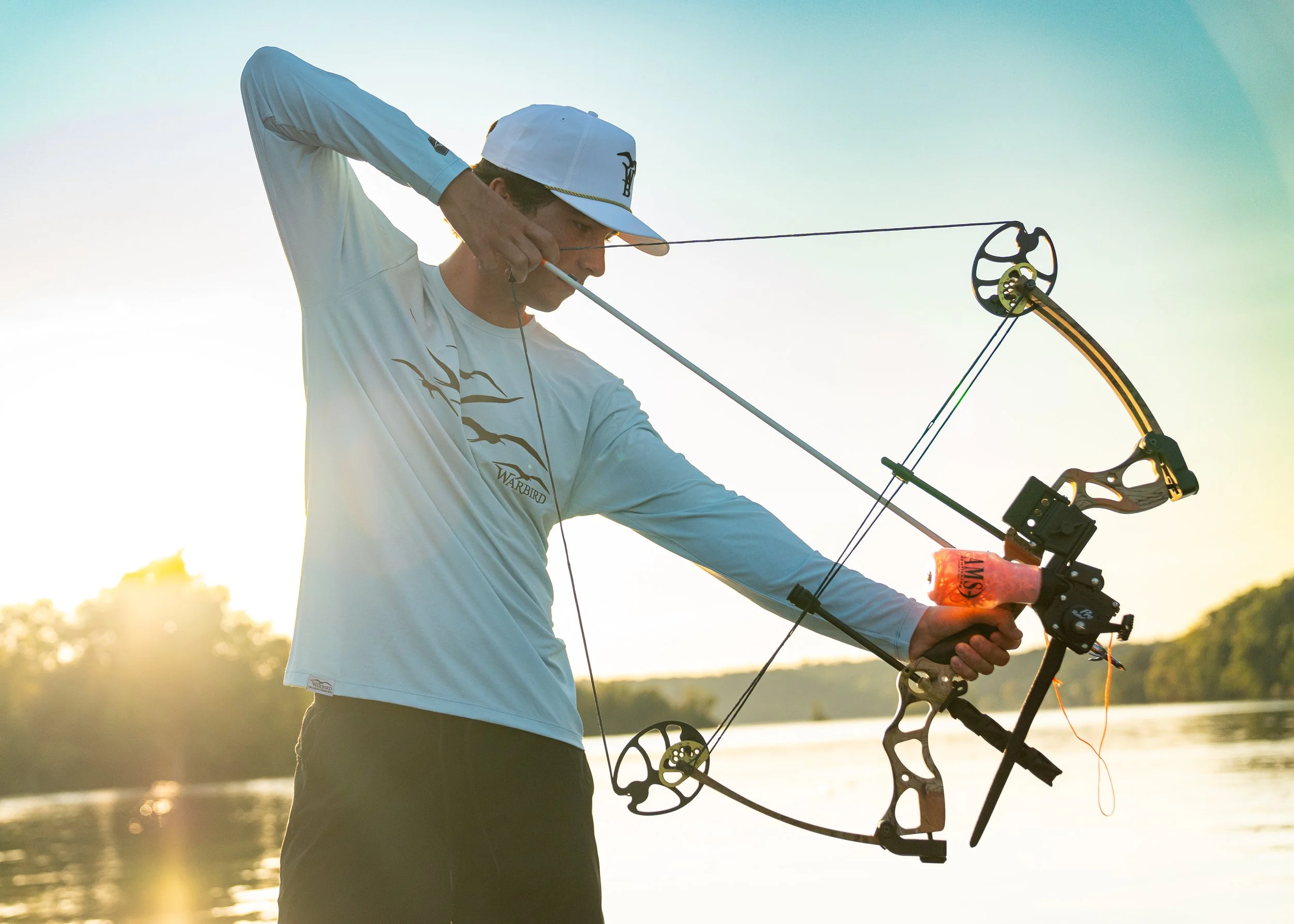 A man is aiming a compound bow near a body of water during sunset or sunrise.