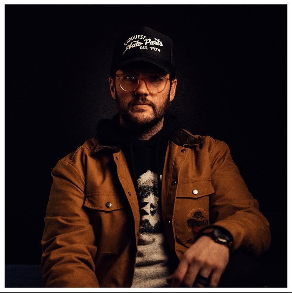 A man wearing glasses, a black baseball cap that says 'Carquest Auto Parts Est. 1974', a brown jacket, and a black hoodie with a face graphic, sitting against a dark background.
