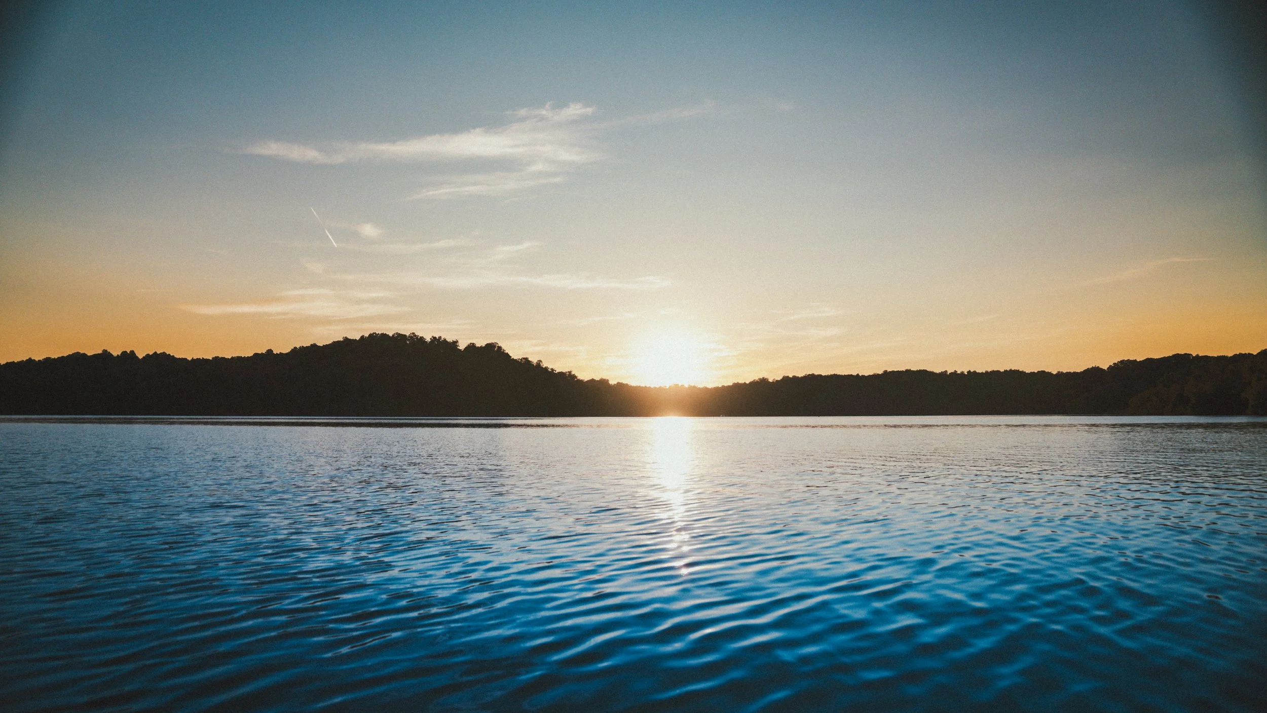 Sunset over a body of water with silhouettes of trees on the horizon.