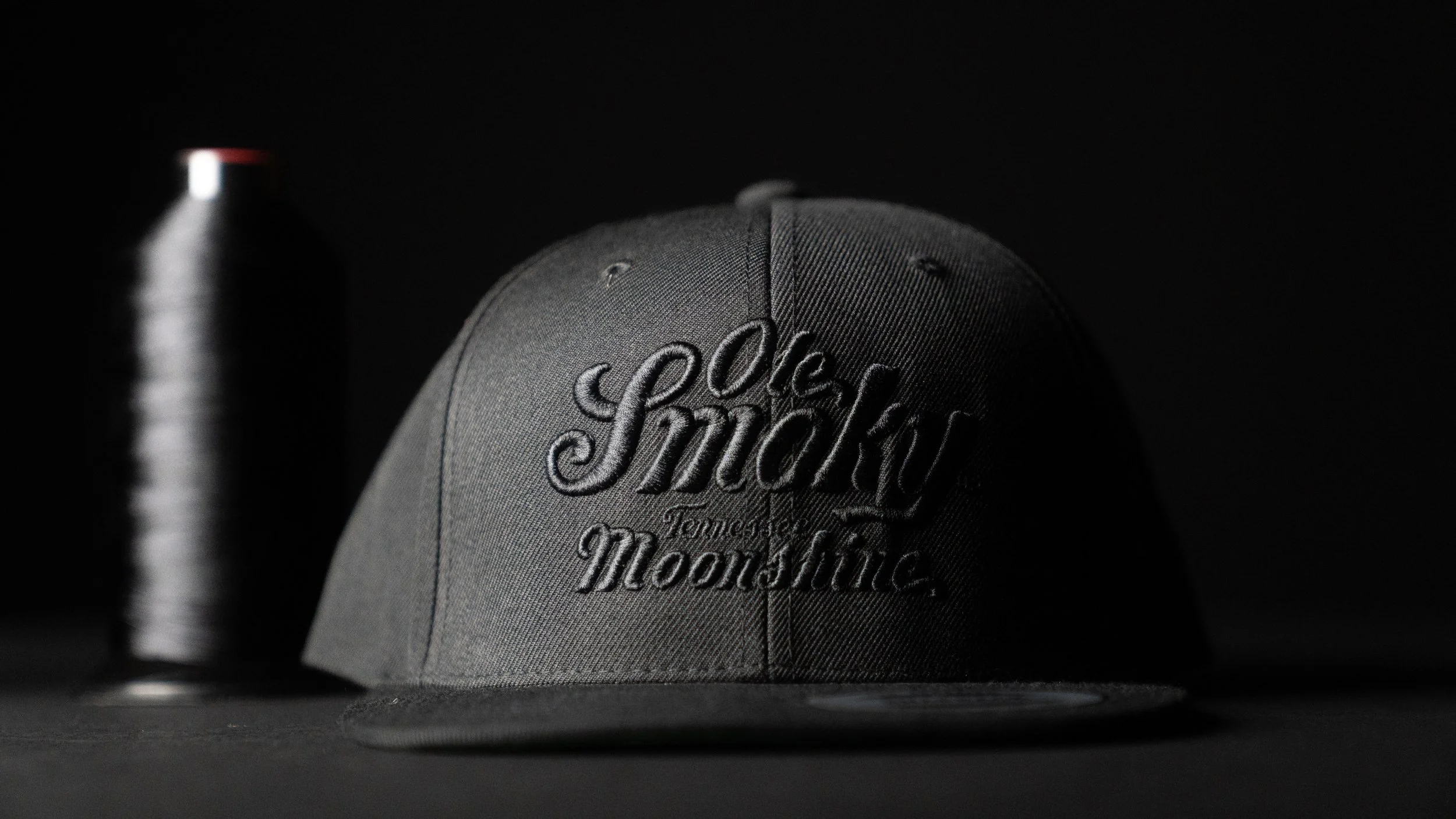 A gray baseball cap with embroidery reading 'Old Smoky Tennessee Moonshine' on the front, placed on a dark surface, with a black water bottle in the background.