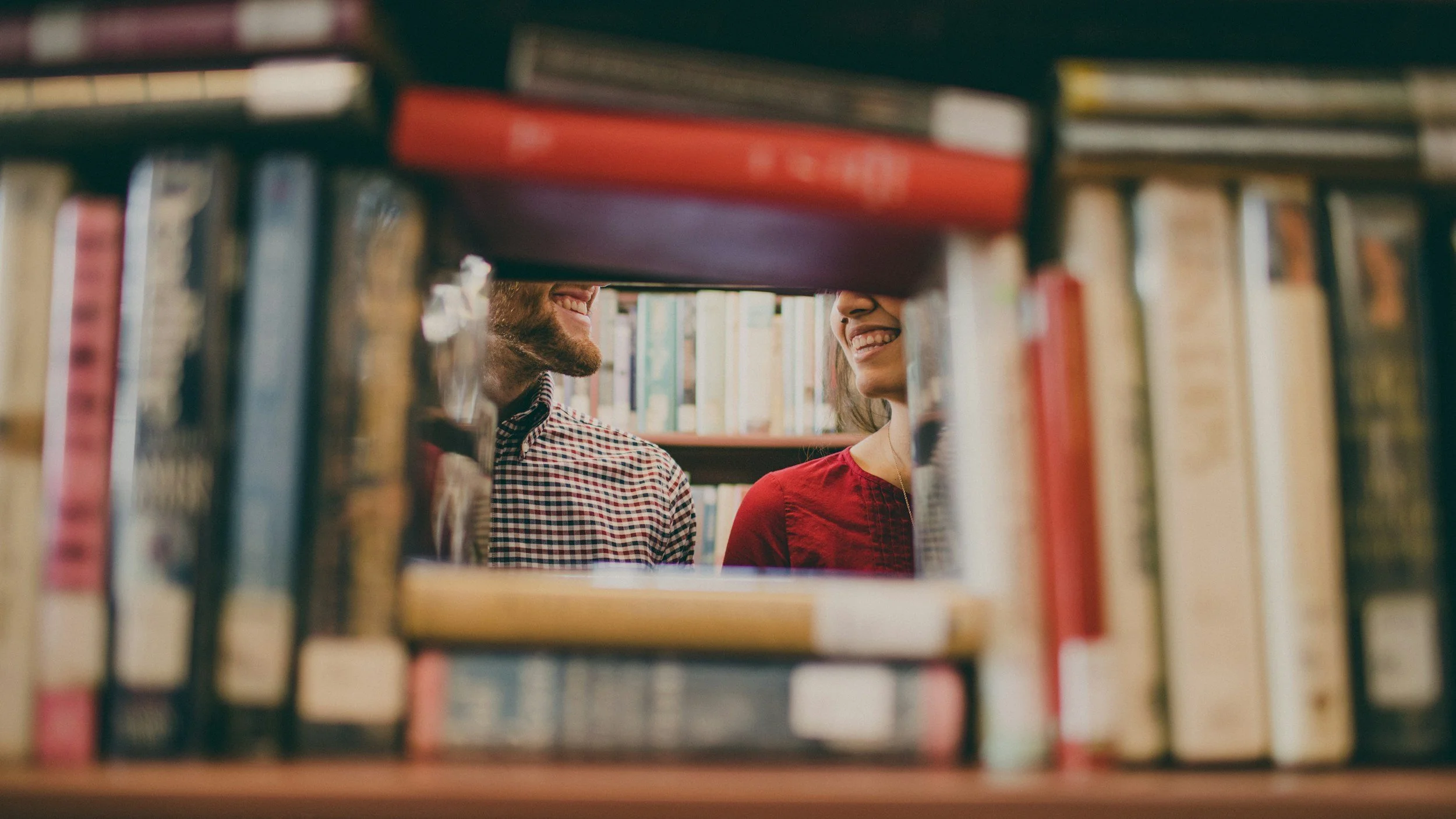 A couple smiling and talking to each other in a library, seen through a gap between books on a shelf.
