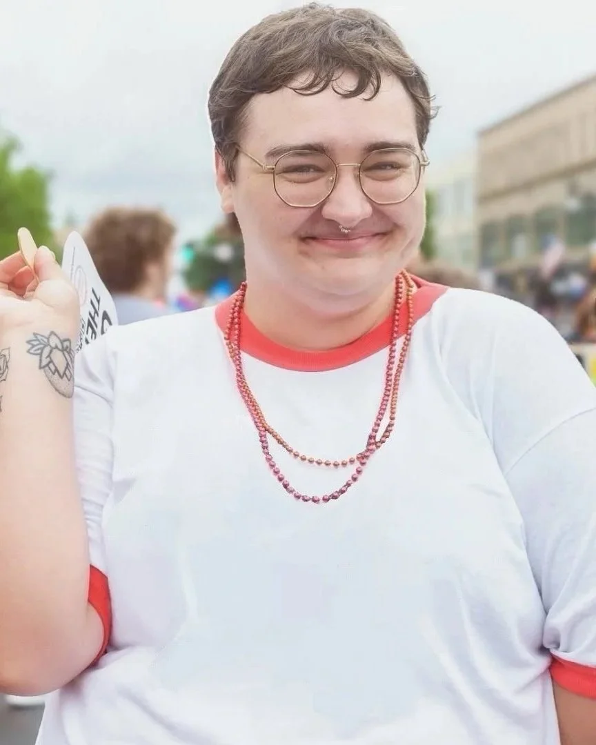 Person with short brown hair, glasses, a septum piercing, and tattoos on their arm, smiling outdoors in a crowd.