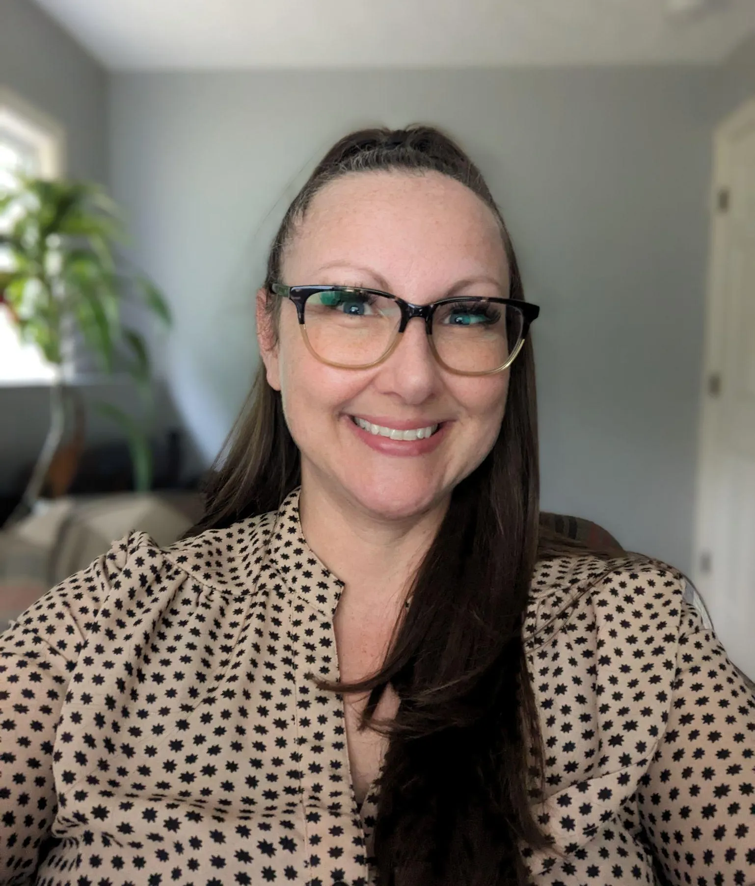 A woman with long brown hair, glasses, and a patterned blouse, smiling for a selfie in a bright room with a plant and a window in the background.