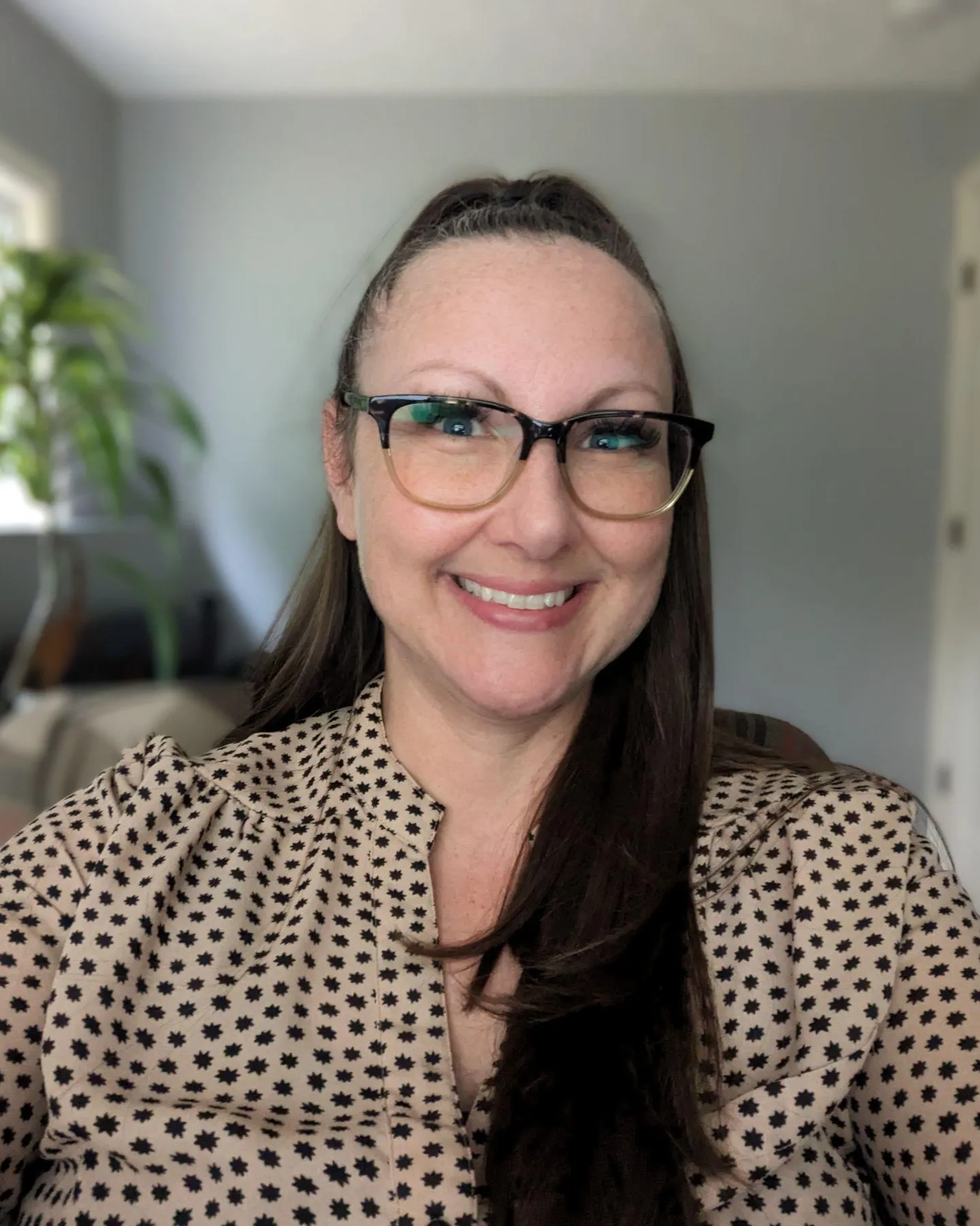 A woman with long dark hair, wearing glasses and a beige blouse with black stars, smiling in a room with gray walls, a window, and a potted plant in the background.