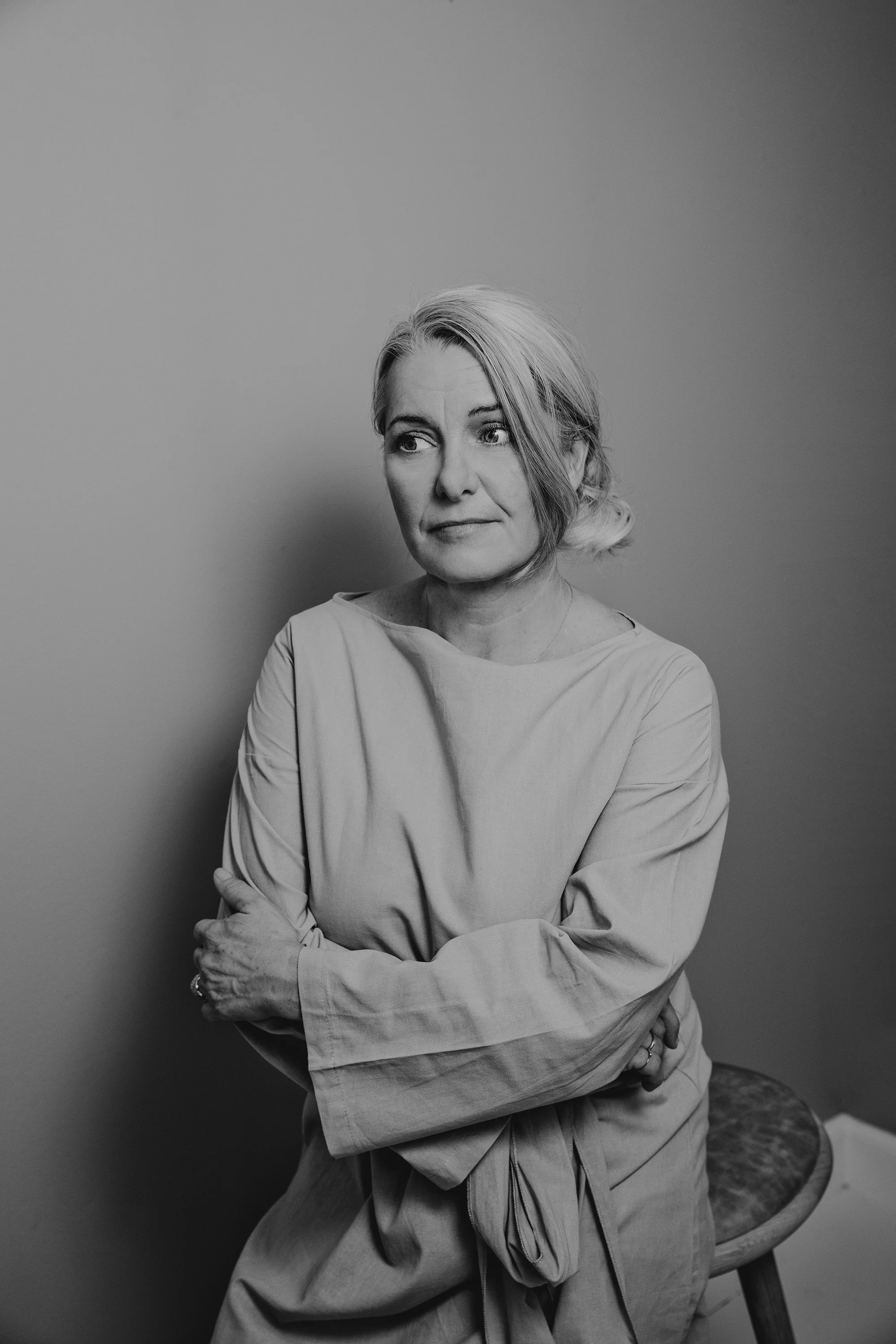 Black and white photo of a woman with short hair sitting on a stool with arms crossed, looking to the side against a plain background.