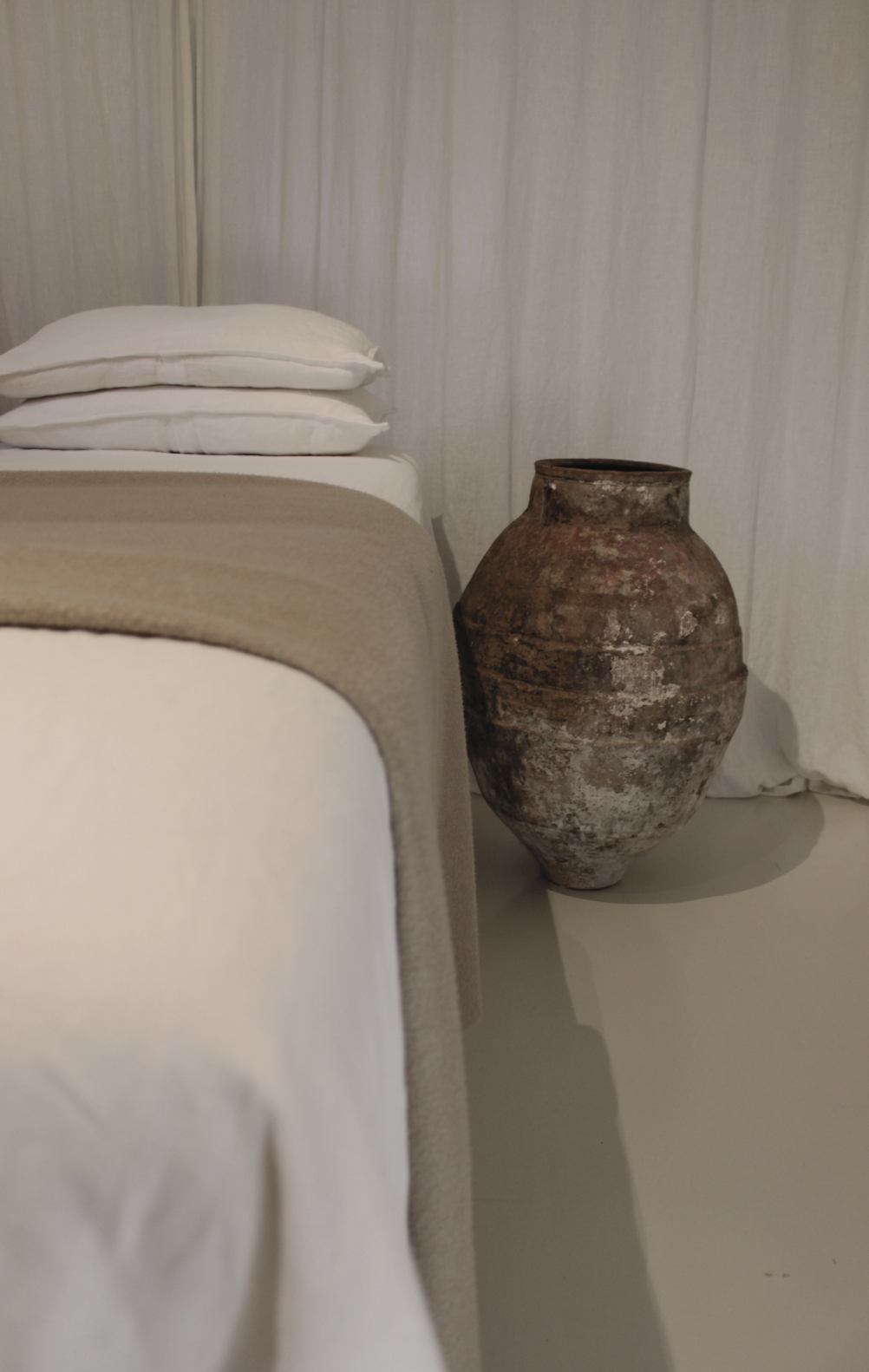 A beige bed with white pillows and a blanket on top of it, next to an ancient, weathered clay urn, with white curtains in the background.