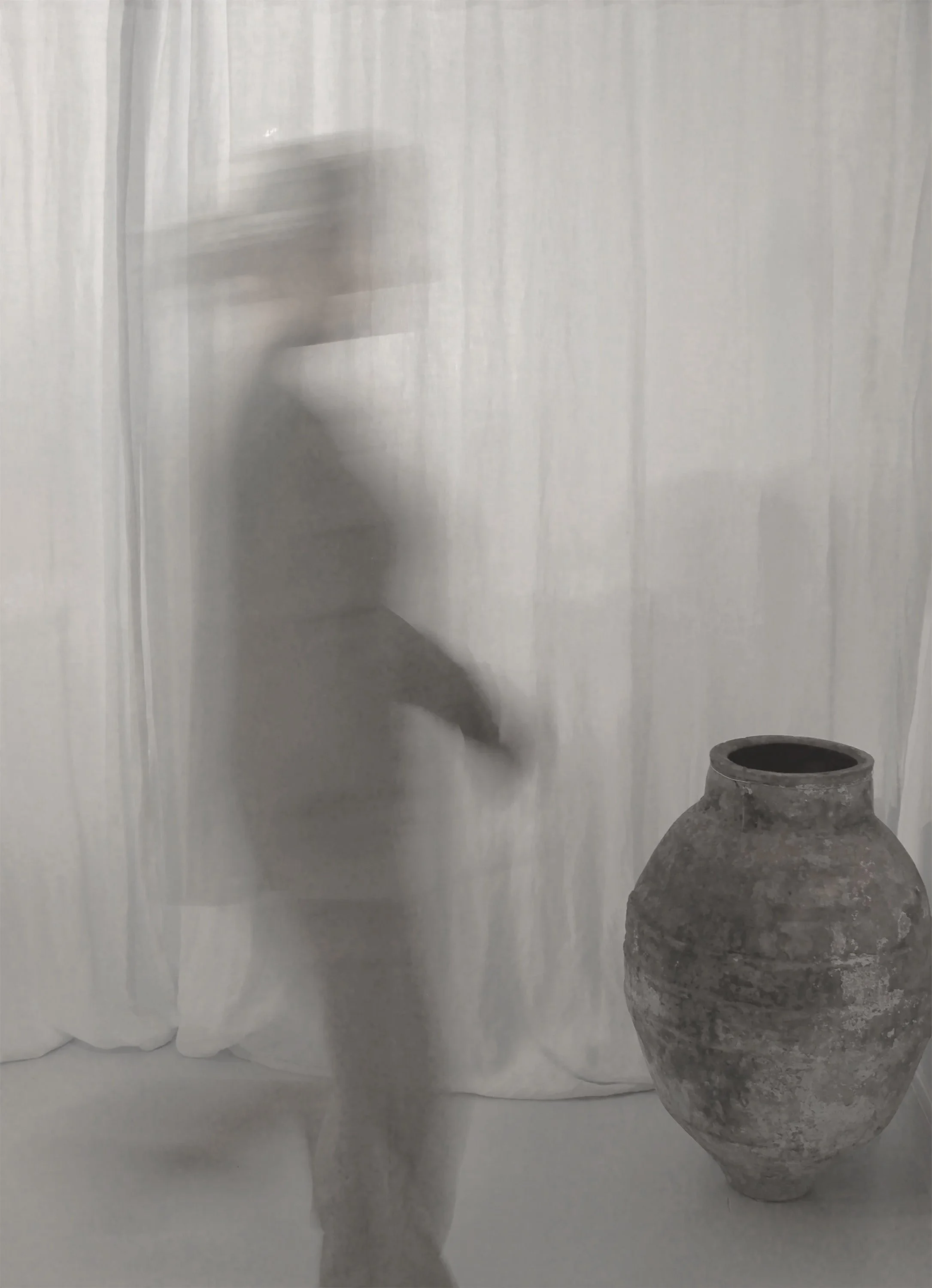 A person blurred in motion standing next to an ancient, large, weathered clay pot against a light-colored curtain background.