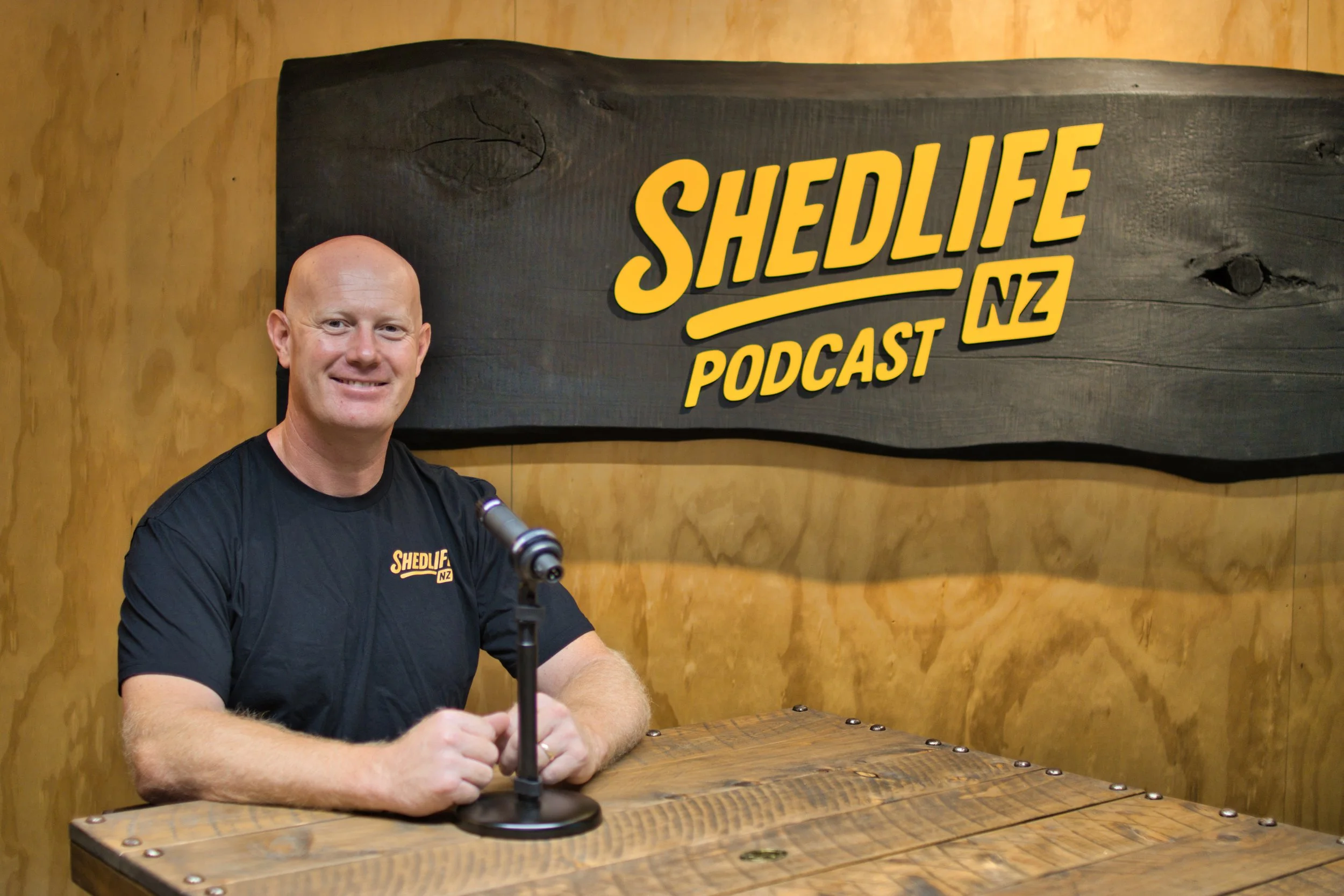 A smiling man with a shaved head, wearing a black T-shirt with a 'Shedlife NZ' logo, sitting at a wooden table with a microphone in front of him. Behind him is a large wooden sign that reads 'Shedlife NZ Podcast,' on a wood-paneled wall.