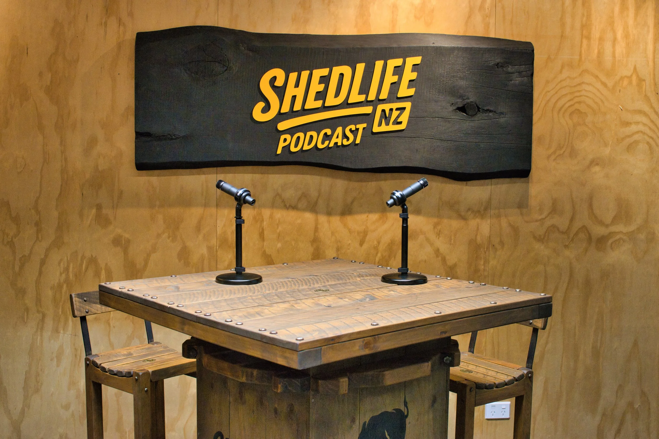 A wooden table with two microphones on stands, set in front of a sign that reads "SHEDLIFE NZ PODCAST" on a wooden wall.