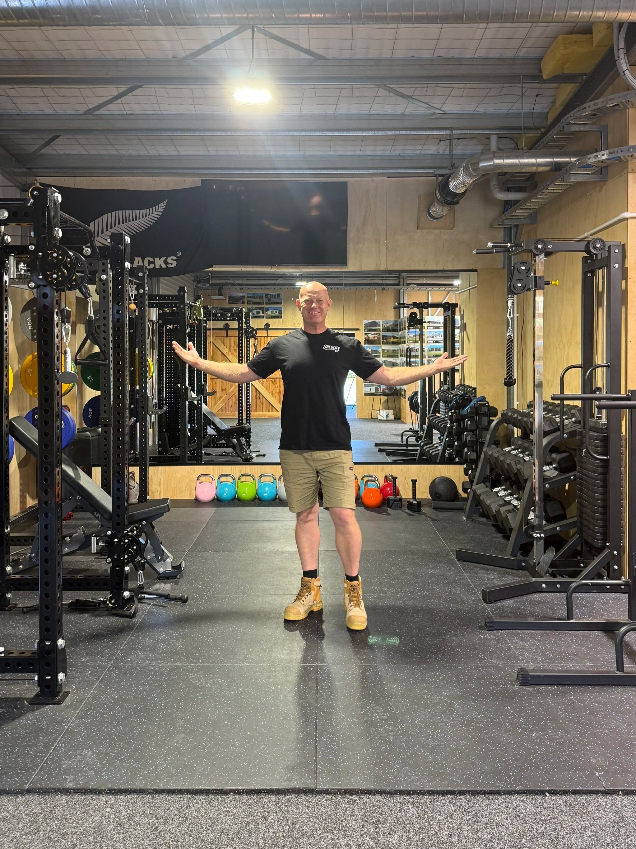 A man (Jason Davis) standing in a gym with his arms outstretched, surrounded by workout equipment including kettlebells, dumbbells, and weight machines, smiling at the camera.