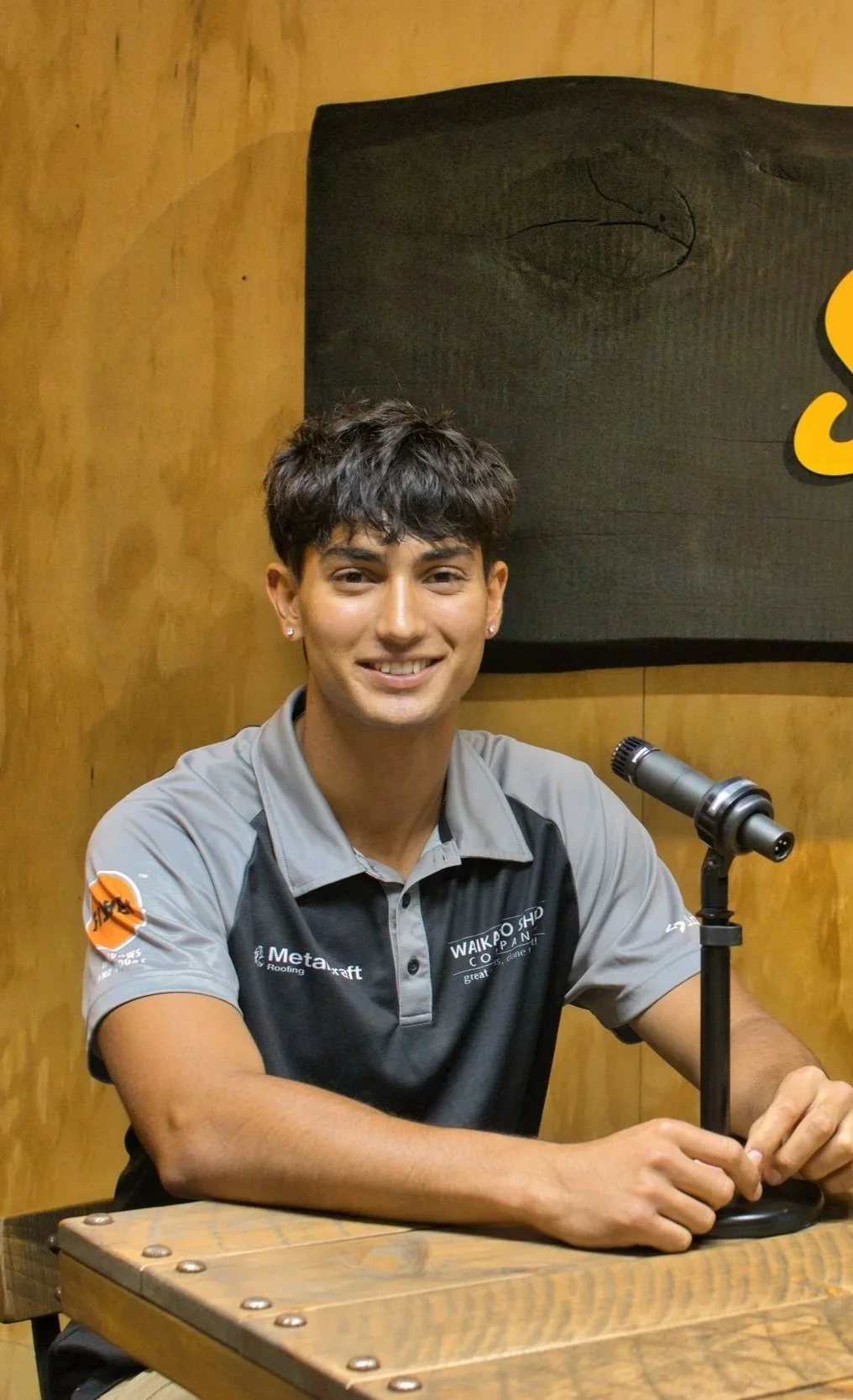 A young man with dark curly hair, wearing earrings and a sports shirt, sitting at a table with a microphone in front of him, smiling at the camera.