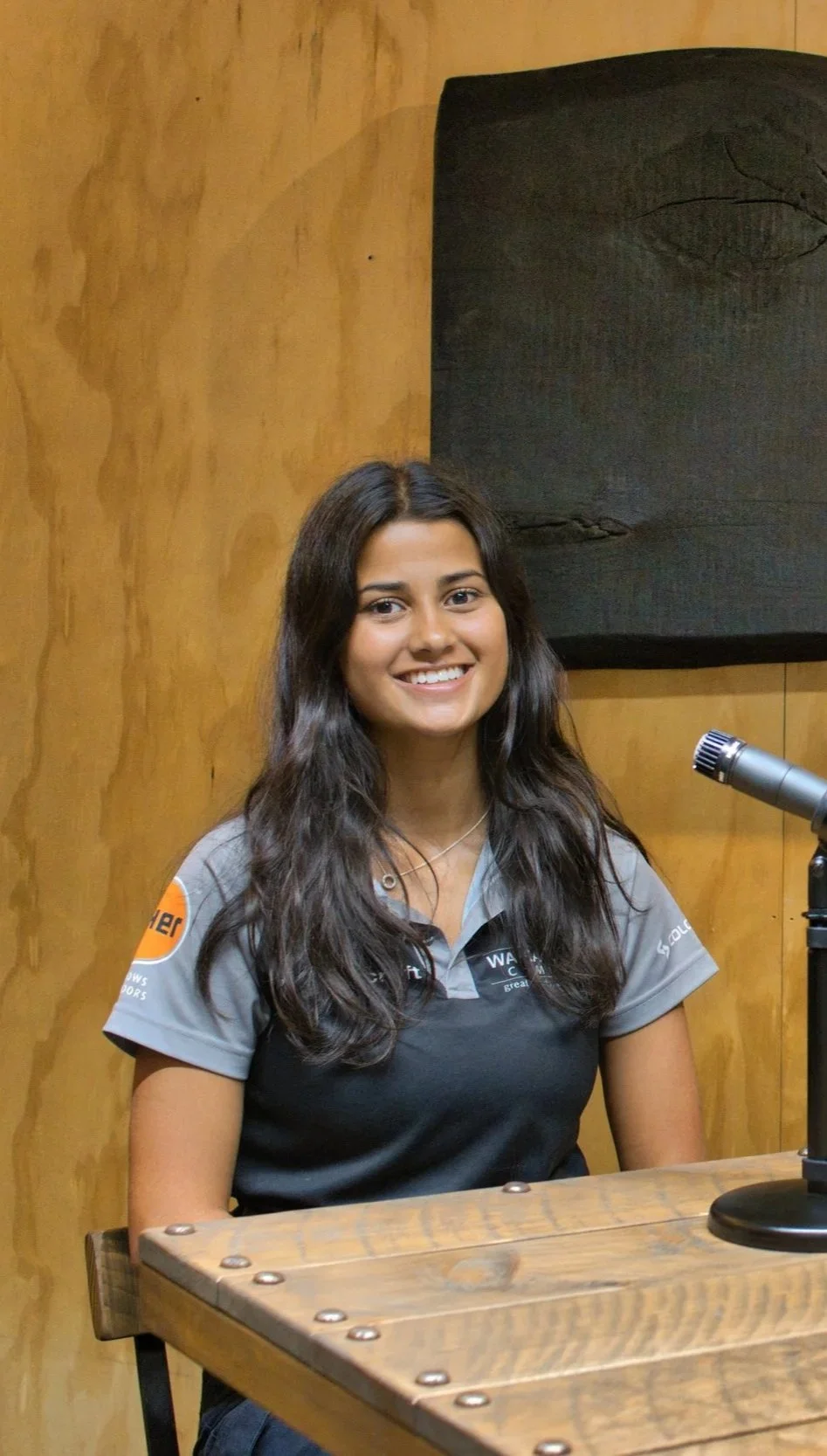 A young woman with long dark hair sitting at a wooden table, smiling, with a microphone nearby, in a room with wooden walls.