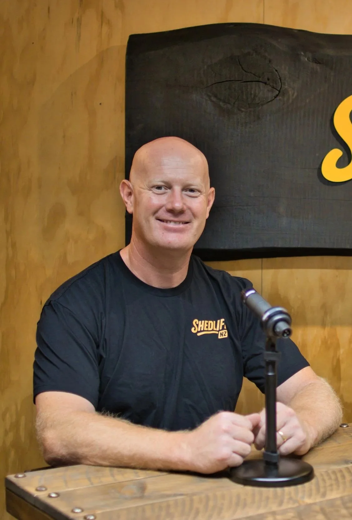 A smiling man in black t-shirt sitting at a wooden table with a microphone in front of him, inside a wood-paneled room.