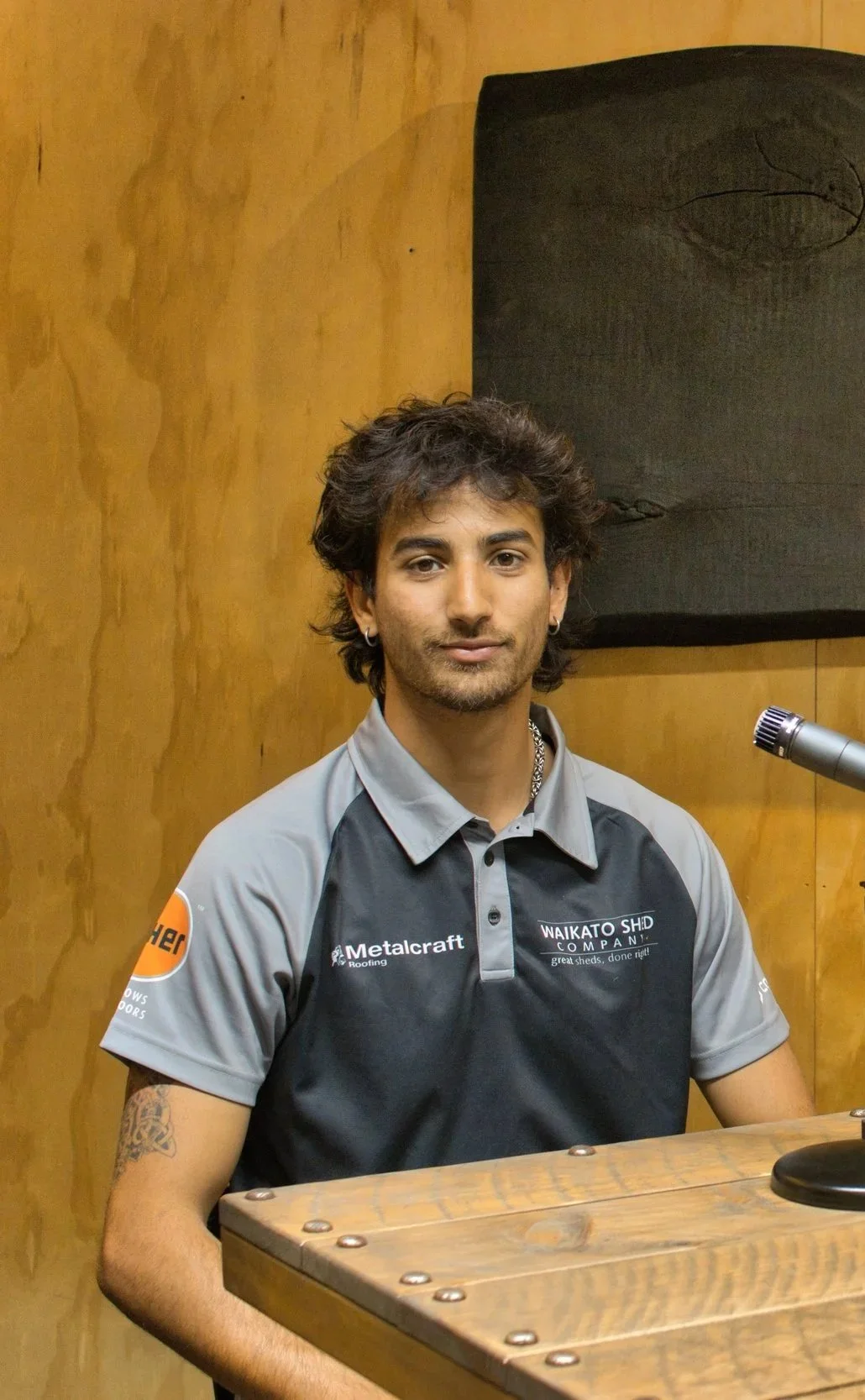 A young man with dark, curly hair and earrings sitting at a wooden table with a microphone nearby, wearing a gray and black sports shirt with various logos, against wooden wall background.