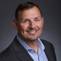 A headshot of a man with short brown hair, smiling, wearing a dark blazer and a blue collared shirt, against a plain white background.