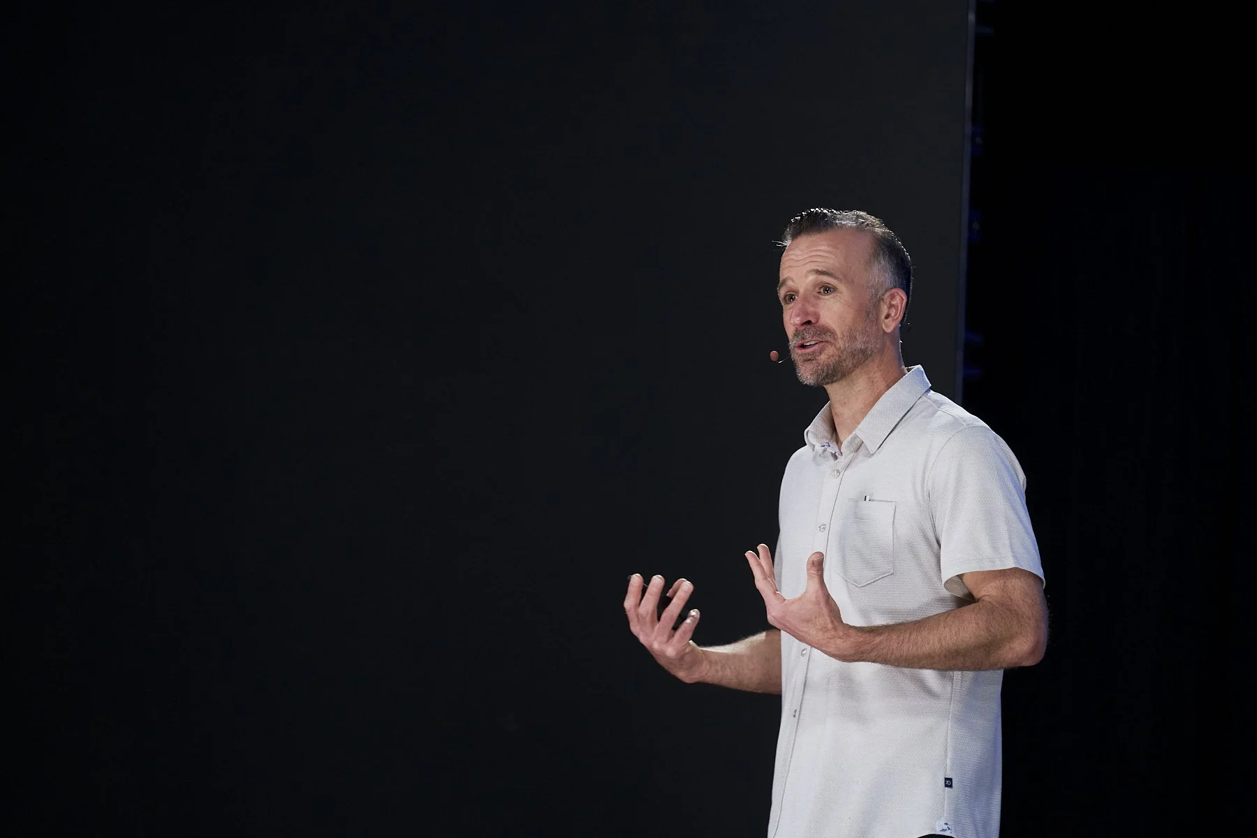 Adam Hill keynote speaking on stage with a microphone headset, gesturing with both hands, against a black background.
