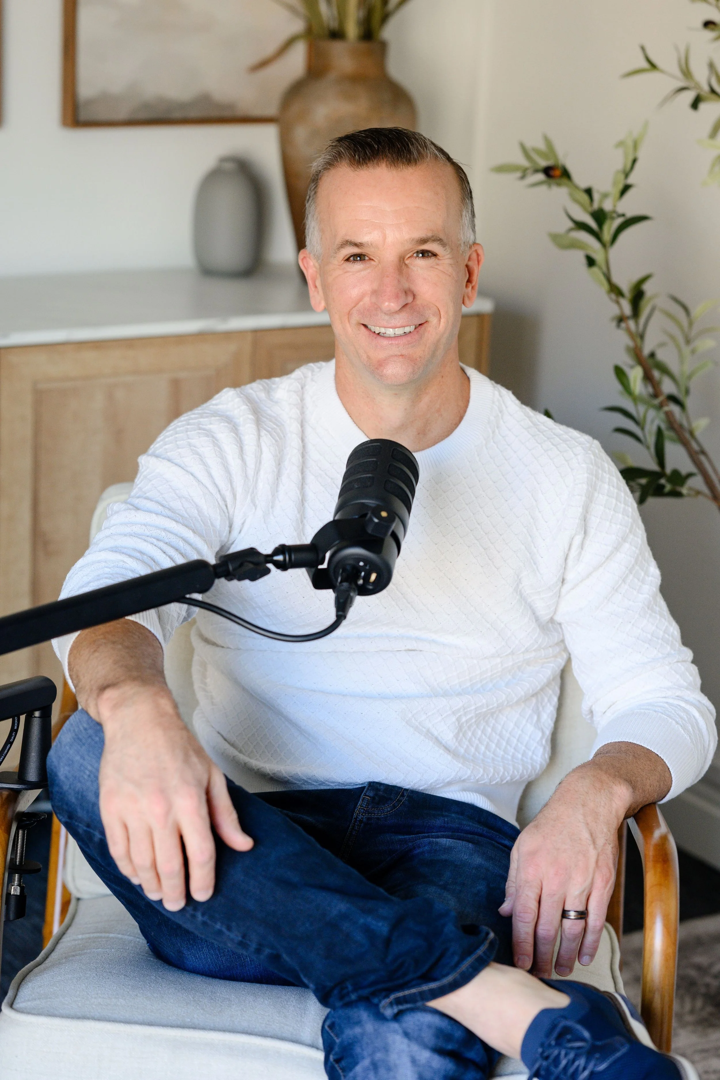 Adam Hill, host of Startup Spouses, wearing a white textured long sleeve shirt, sitting in a chair with crossed legs, and speaking into a black microphone.