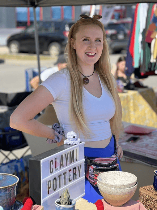 A young woman with long blonde hair, smiling, standing at a pottery booth at an outdoor market. She is wearing a white crop top, a necklace, sunglasses on her head, and a scrunchie on her wrist. The booth has pottery items, and a sign that reads 'Claymy Pottery'.