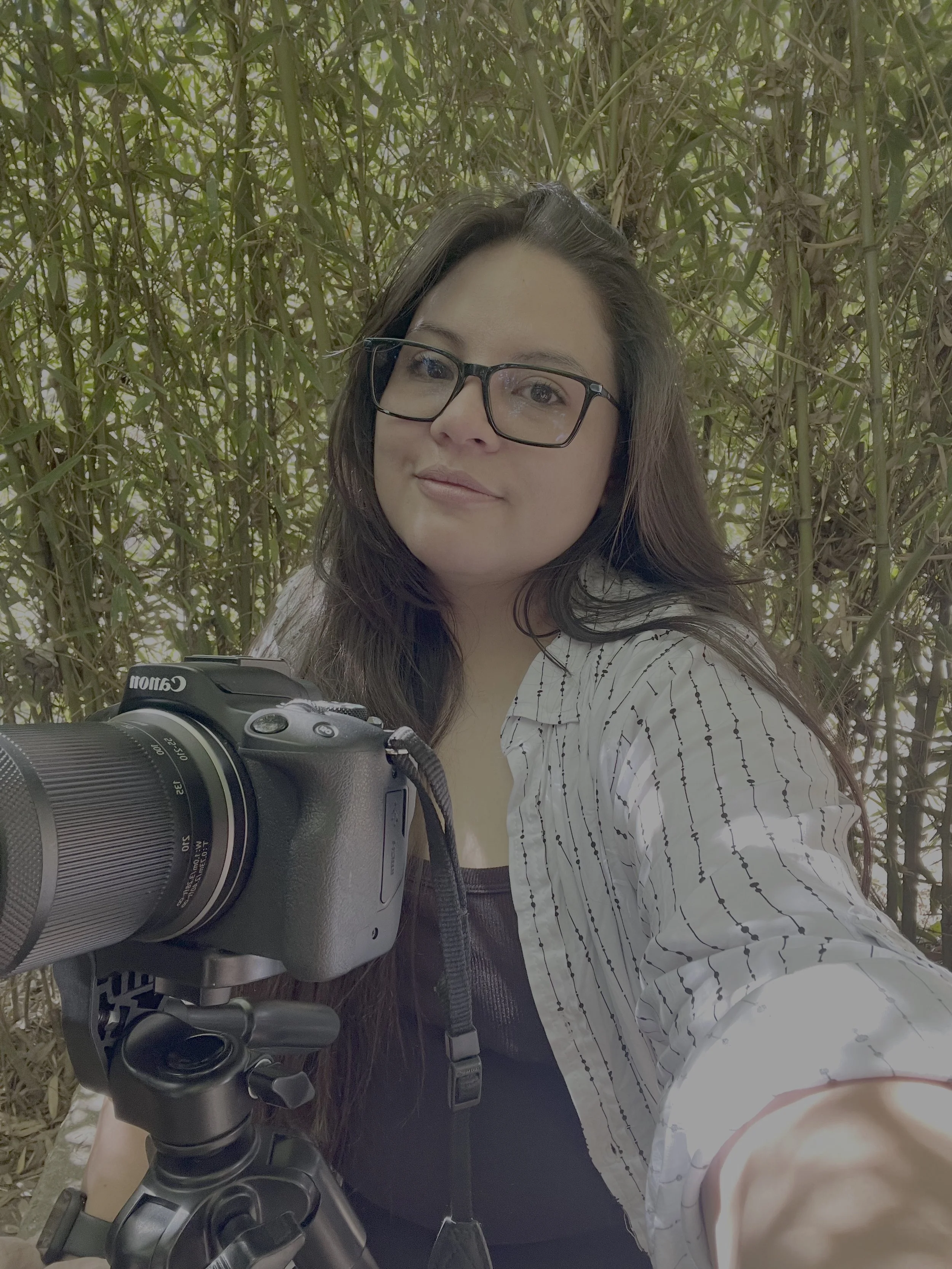 A woman wearing glasses taking a selfie in front of dense green foliage while holding a camera on a tripod.