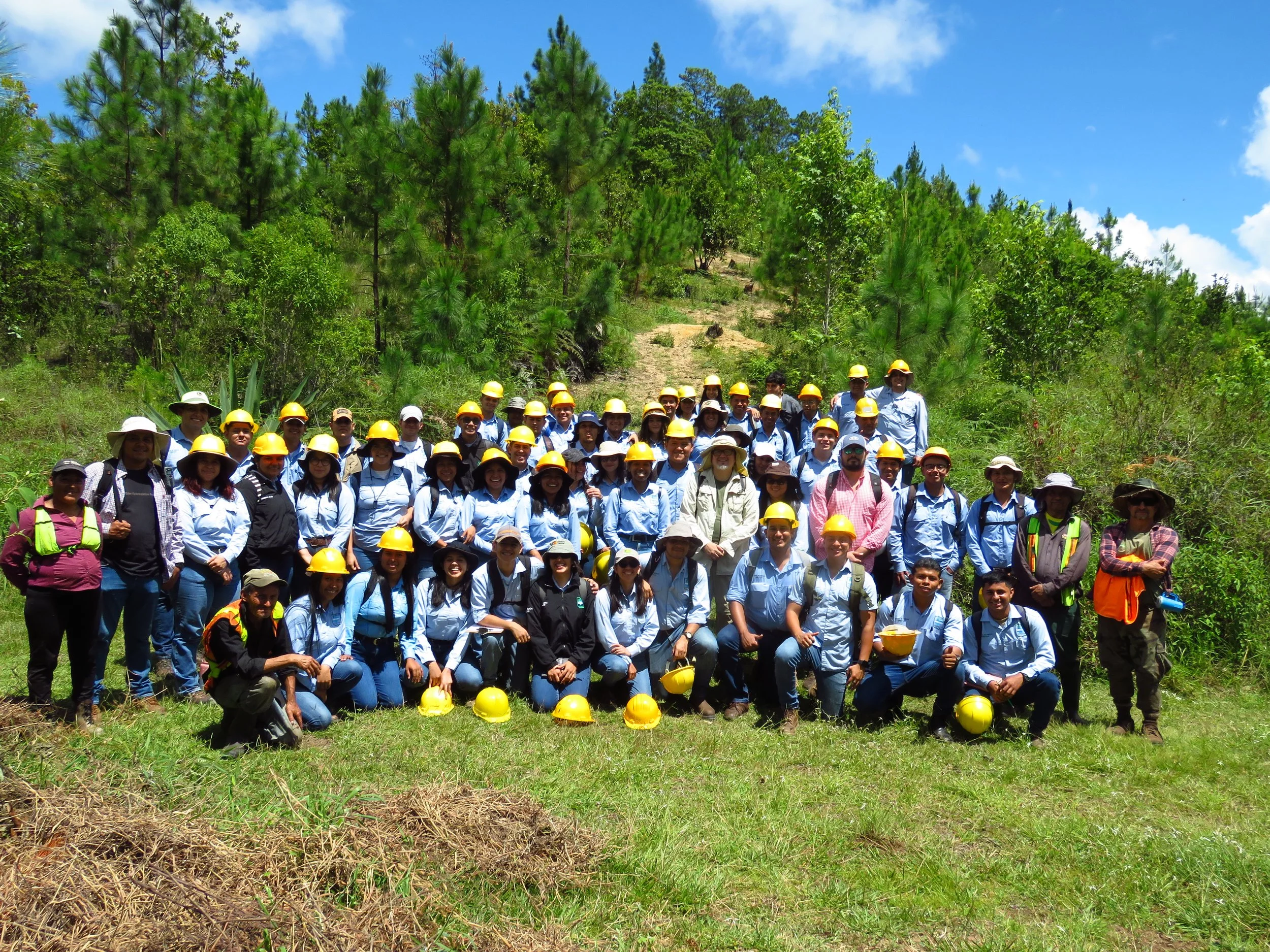 Group of people outdoors in a wooded area, wearing hats and safety vests, posing for a photo.