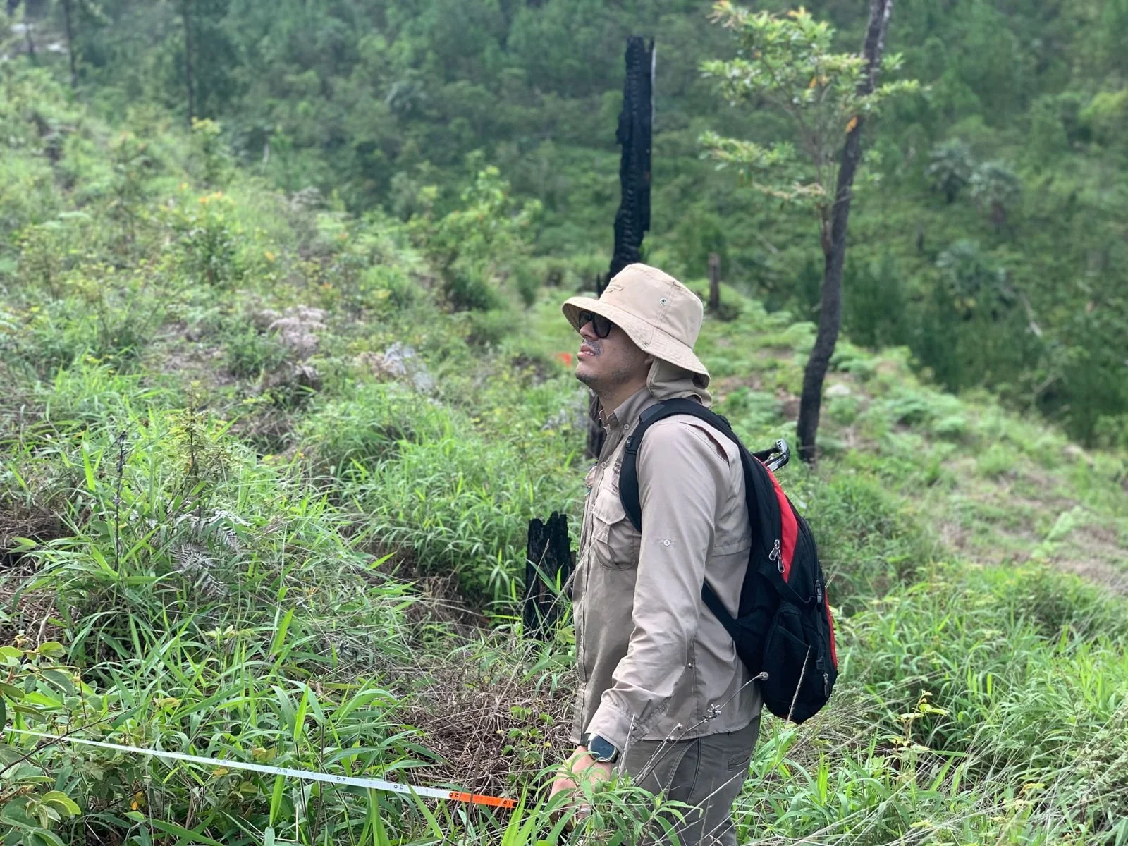 A man wearing a beige hat, sunglasses, a beige long-sleeve shirt, beige pants, and a black backpack stands in a green forested area with tall grass and trees.