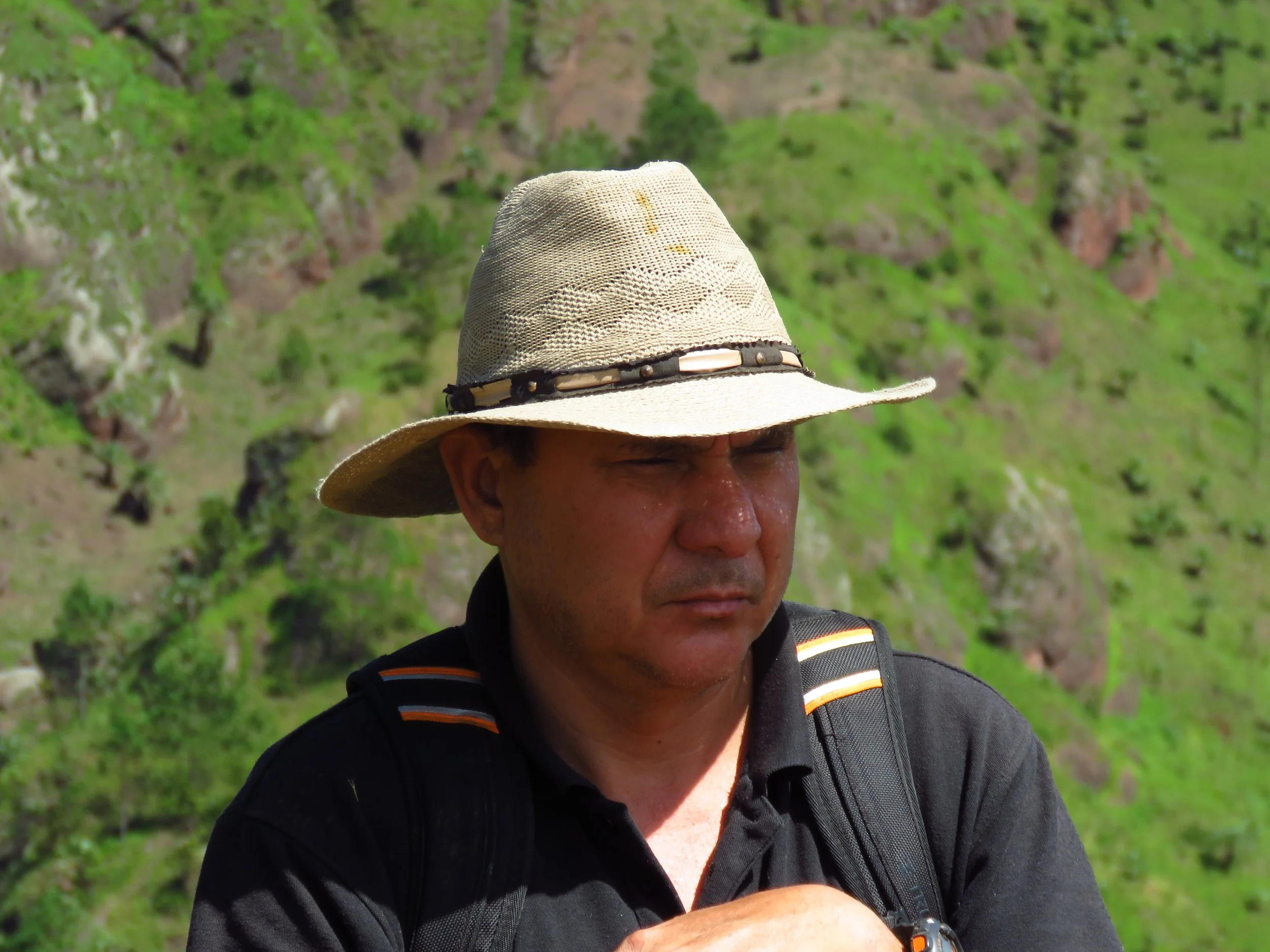 A man with tan skin wearing a wide-brimmed hat and a black shirt, carrying a backpack, outdoors with green hills in the background.