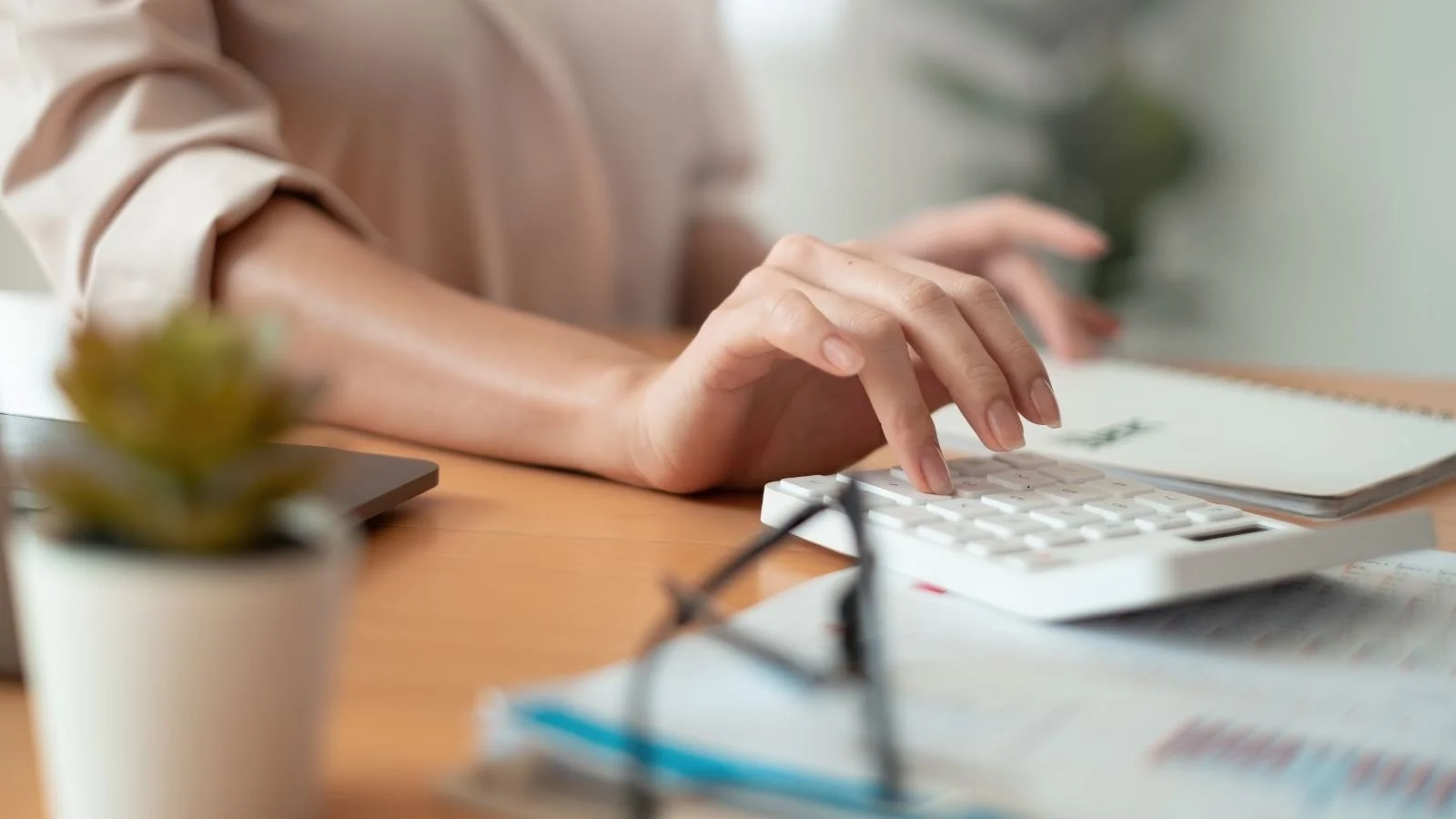 Person typing on a calculator at a desk with glasses, a notebook, a plant, and papers.