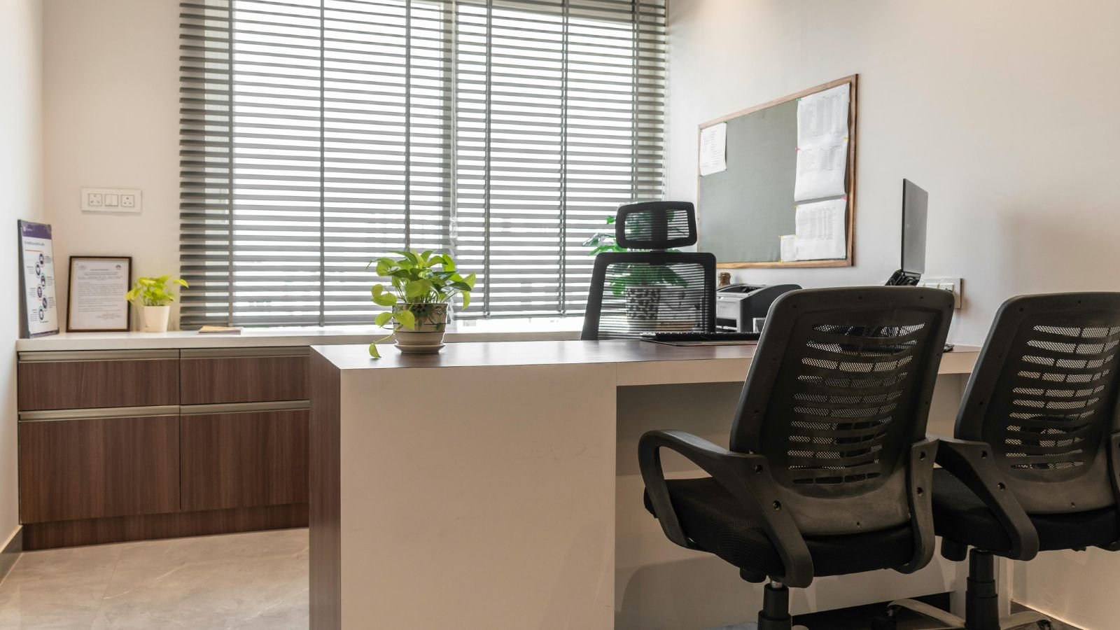 An empty modern office with a desk, black office chairs, a computer, potted plants, a window with horizontal blinds, and a bulletin board on the wall.