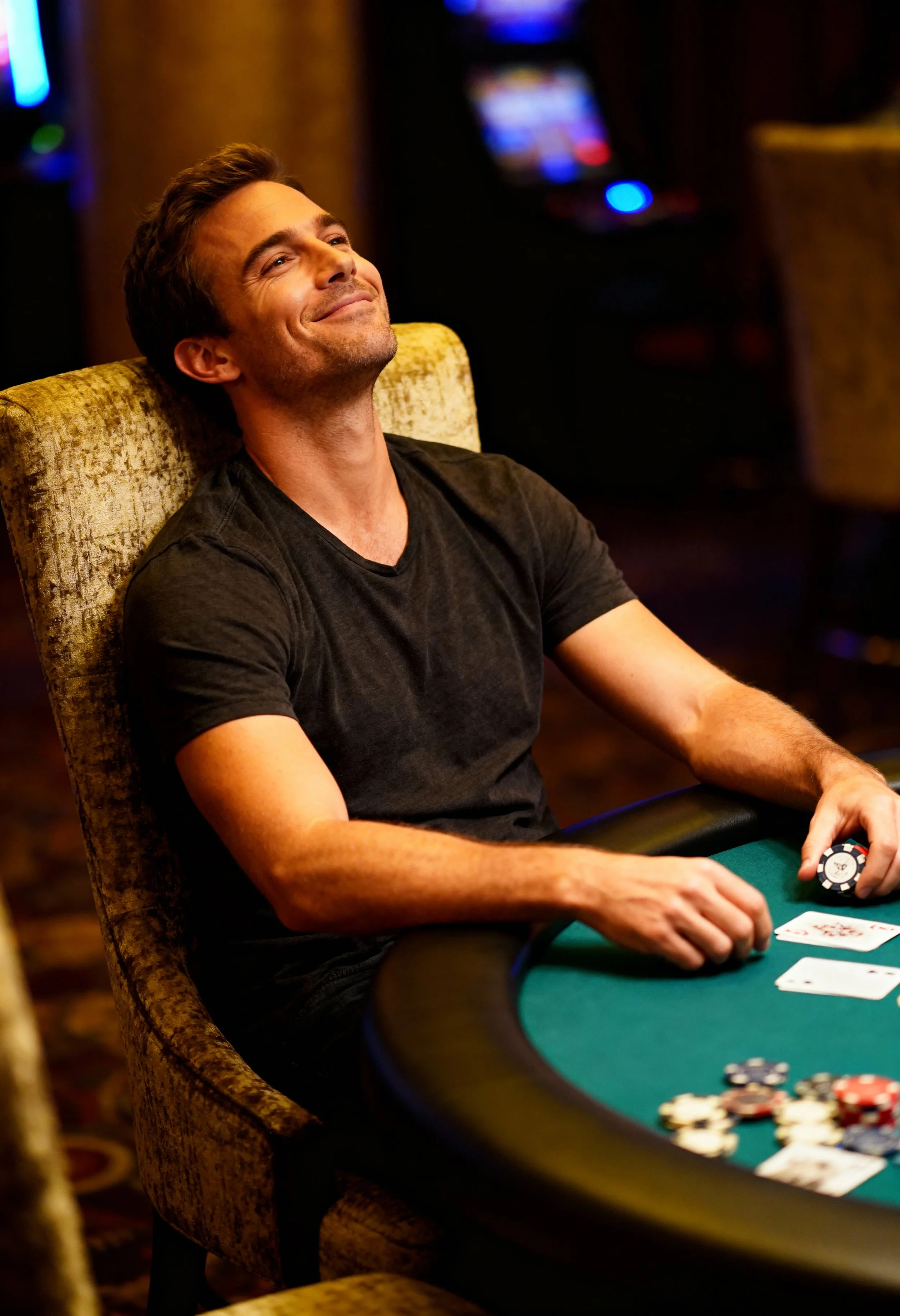 A man smiling and relaxing at a poker table in a casino, holding poker chips with cards in front of him.