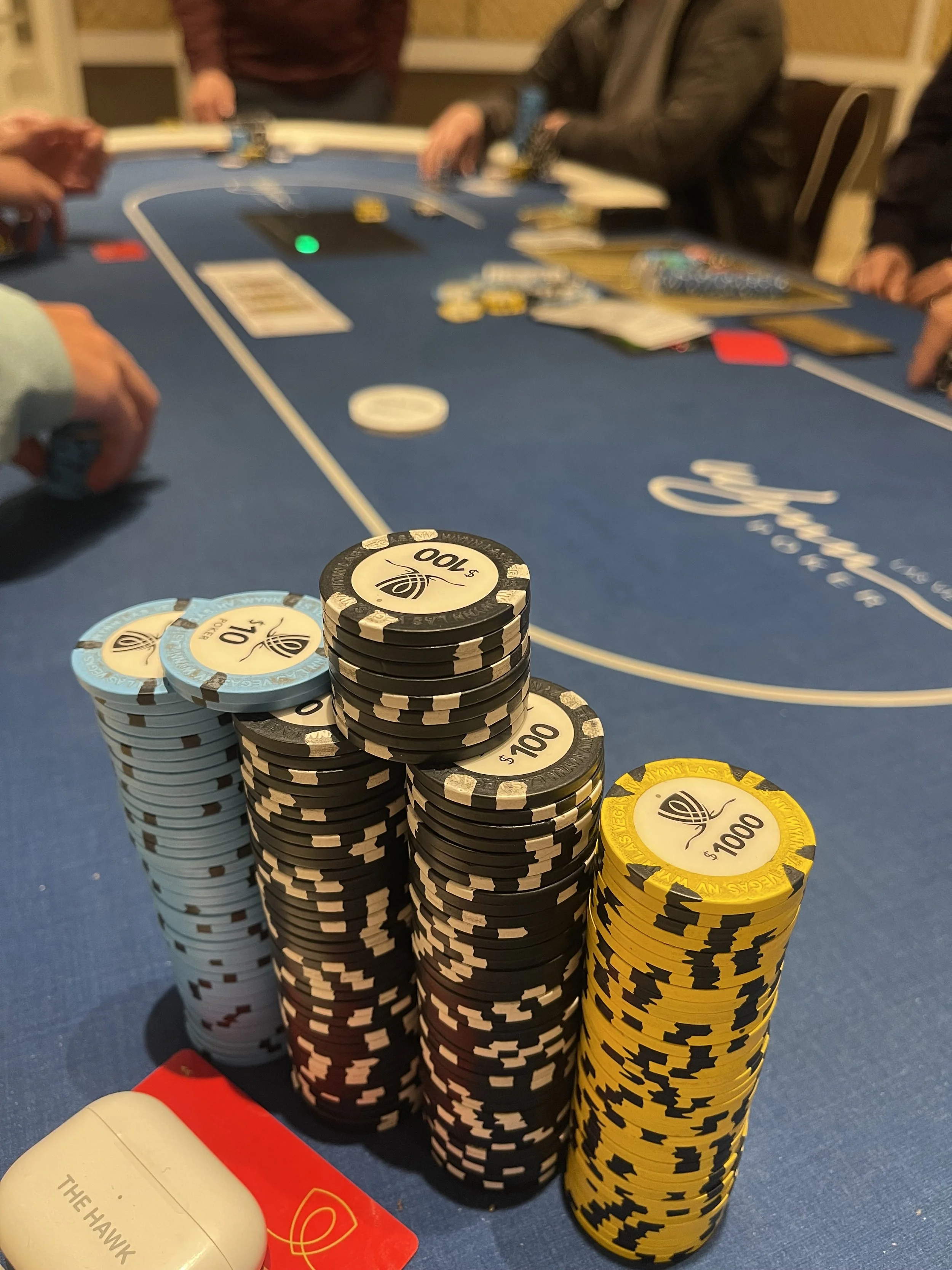 Stacks of poker chips in black, blue, and yellow colors on a blue poker table, with people playing poker in the background.