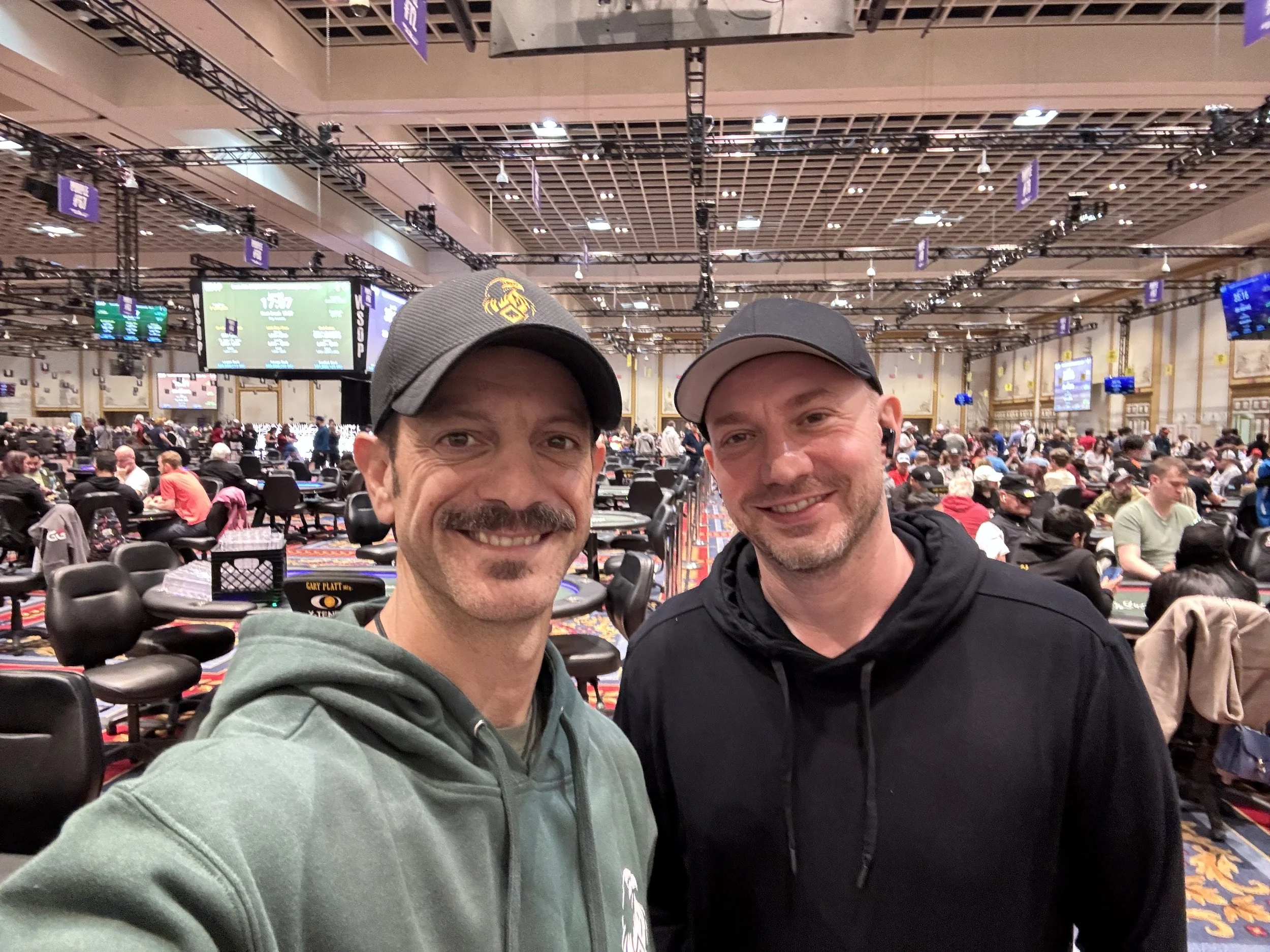 David Richards & Sasha | Poker with a Purpose Two men taking a selfie at a large poker tournament, surrounded by many players seated at poker tables in a spacious, well-lit casino hall.