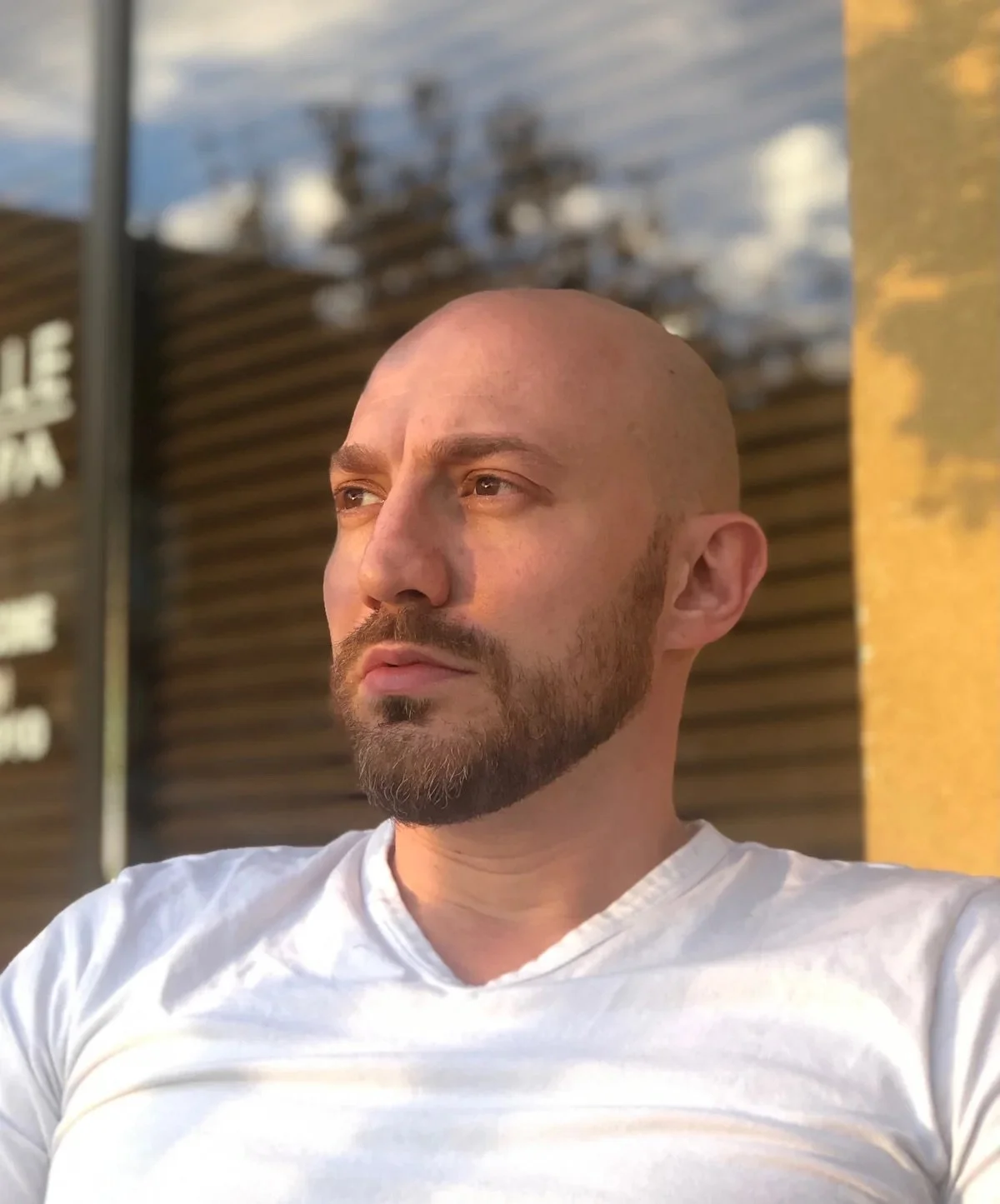 Sasha with a beard and mustache, wearing a white shirt, looking off into the distance outdoors.
