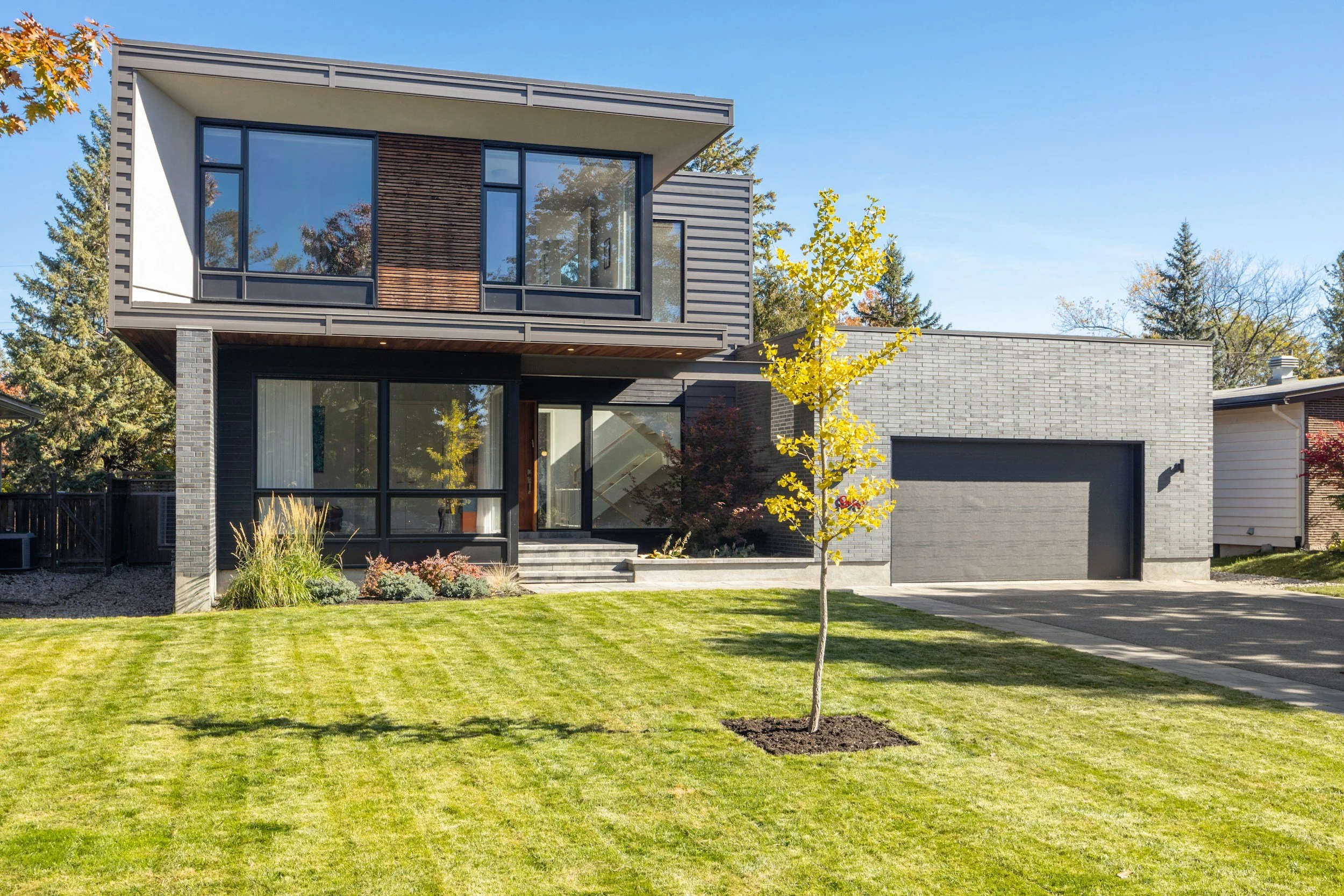 Modern two-story house with dark brick and siding, large windows, and a garage. Green lawn with a small tree in the front yard.