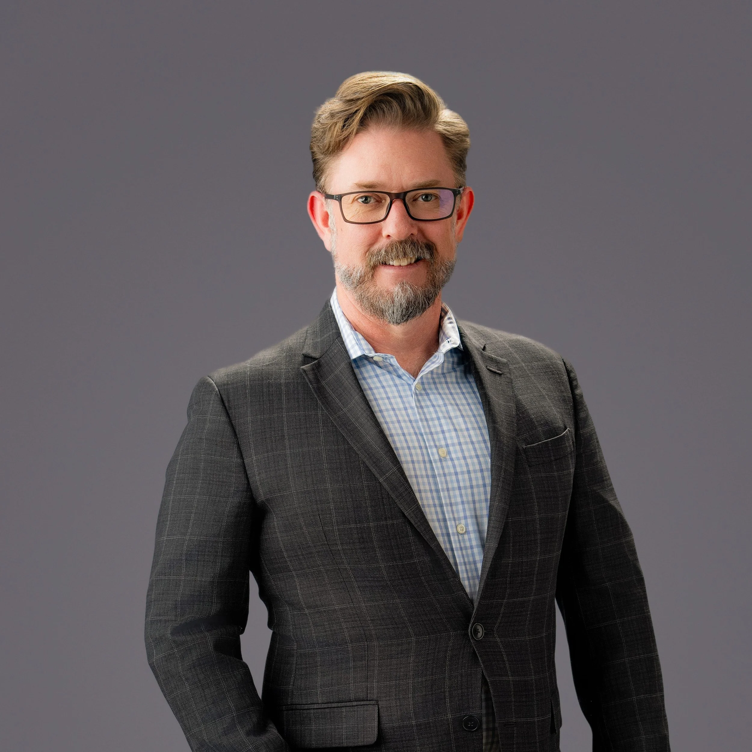 A middle-aged man with glasses and a beard, wearing a checkered shirt and a dark blazer, standing against a plain gray background.