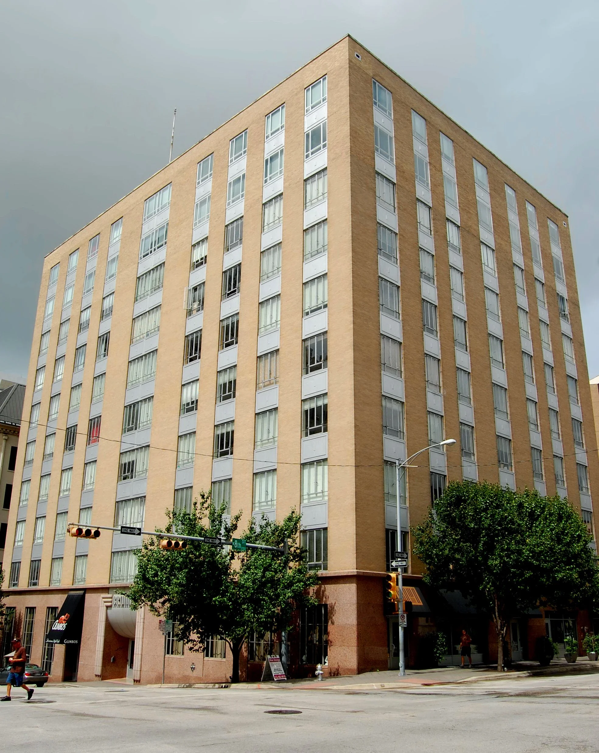 A tall, beige multi-story building on a city street with cars, trees, and pedestrians, overcast sky.