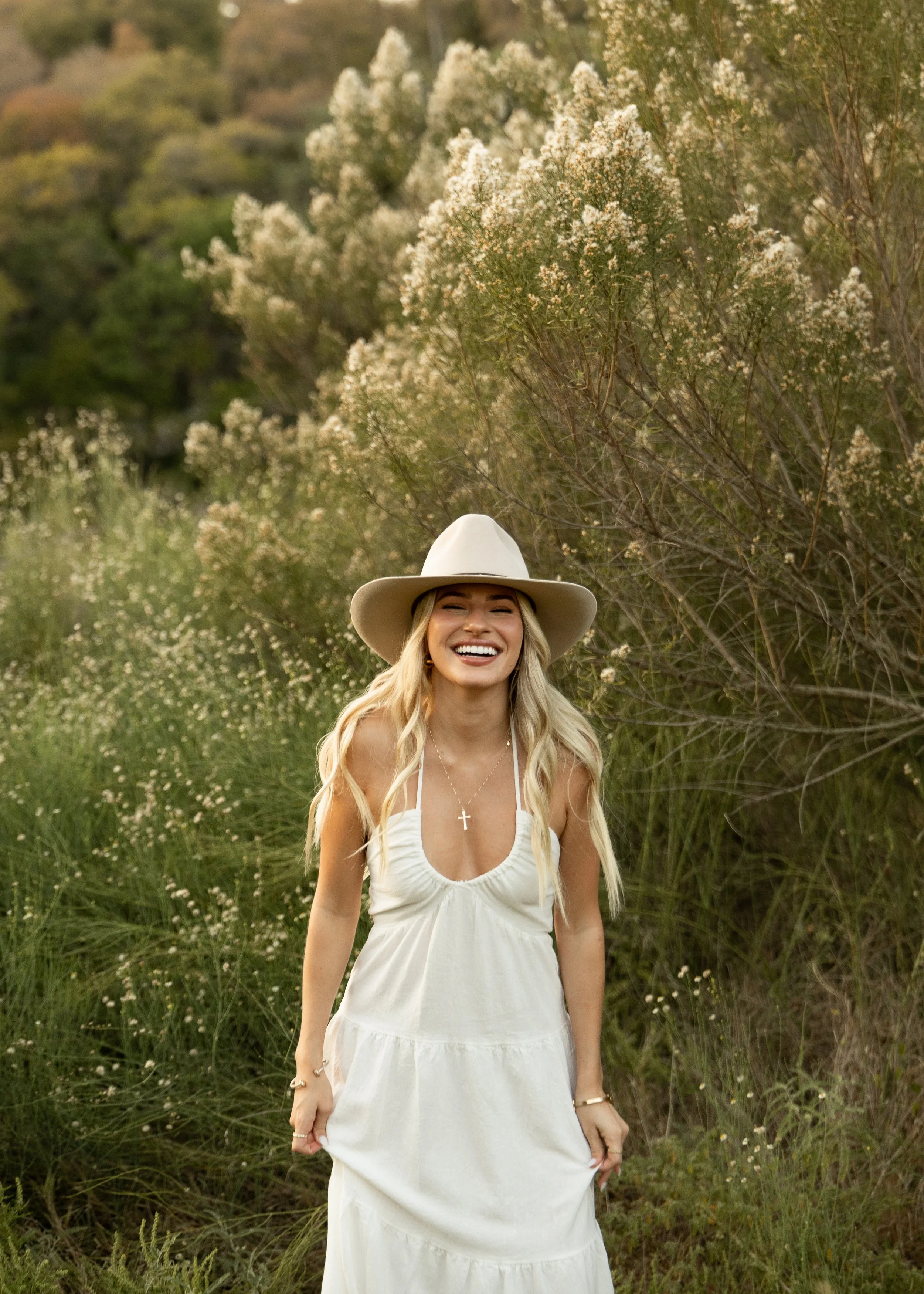 A woman in a white dress and large hat smiling outdoors near greenery and flowering bushes.