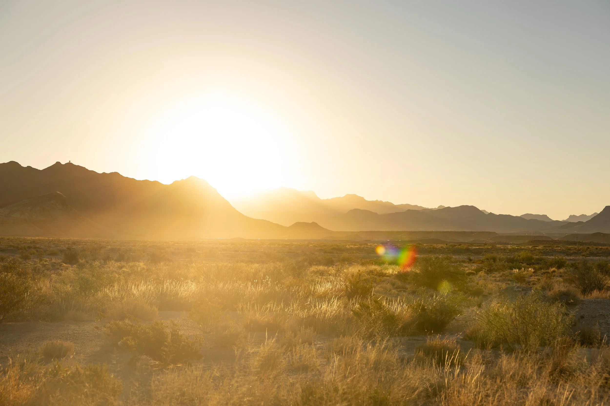 Desert landscape with mountains at sunset, featuring dry vegetation and a bright, setting sun.