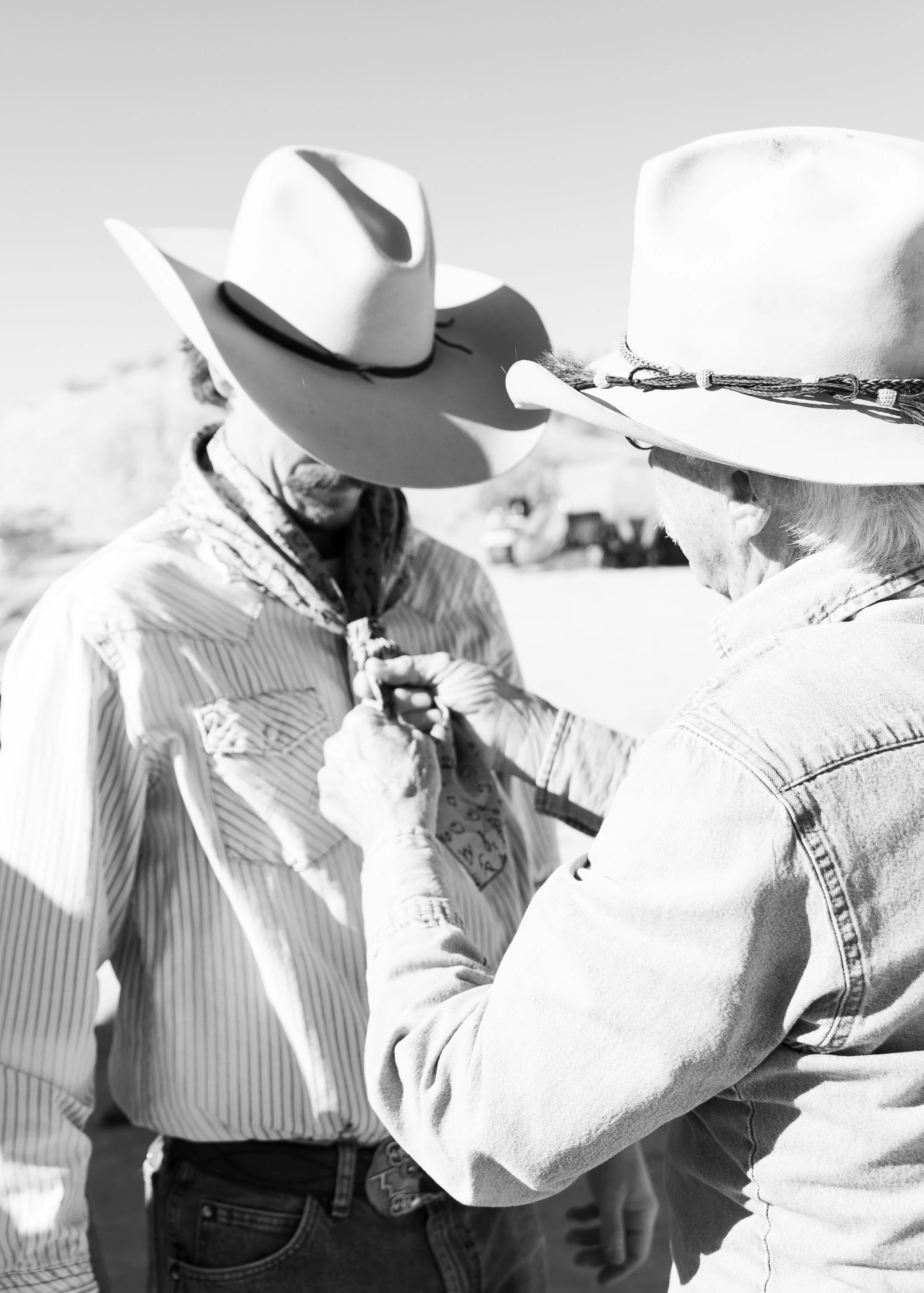 Two people wearing cowboy hats, one helping the other tie a bandana around his neck outdoors.