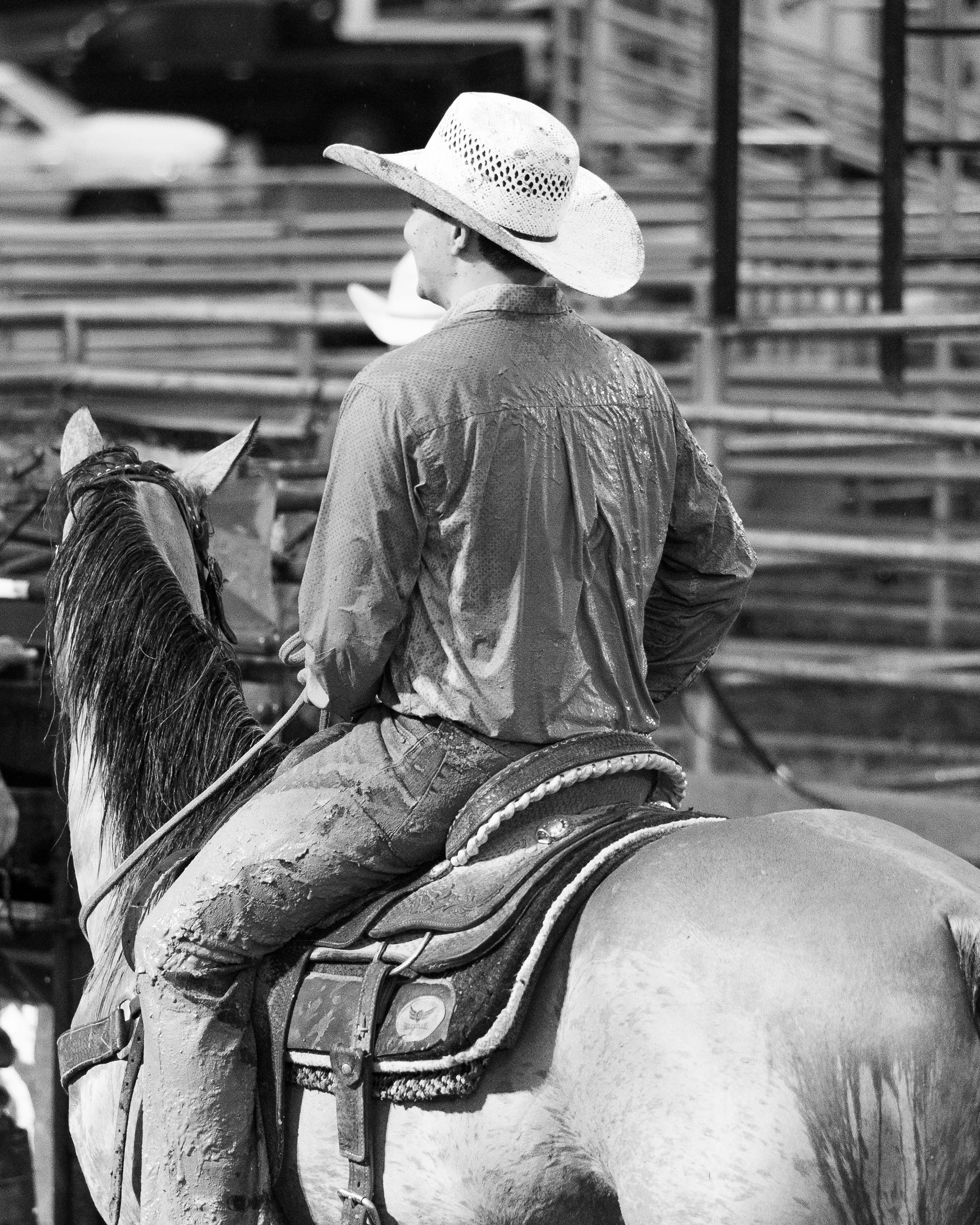 A person wearing a wide-brimmed straw hat and a wet shirt sits on a horse, with wooden bleachers in the background.