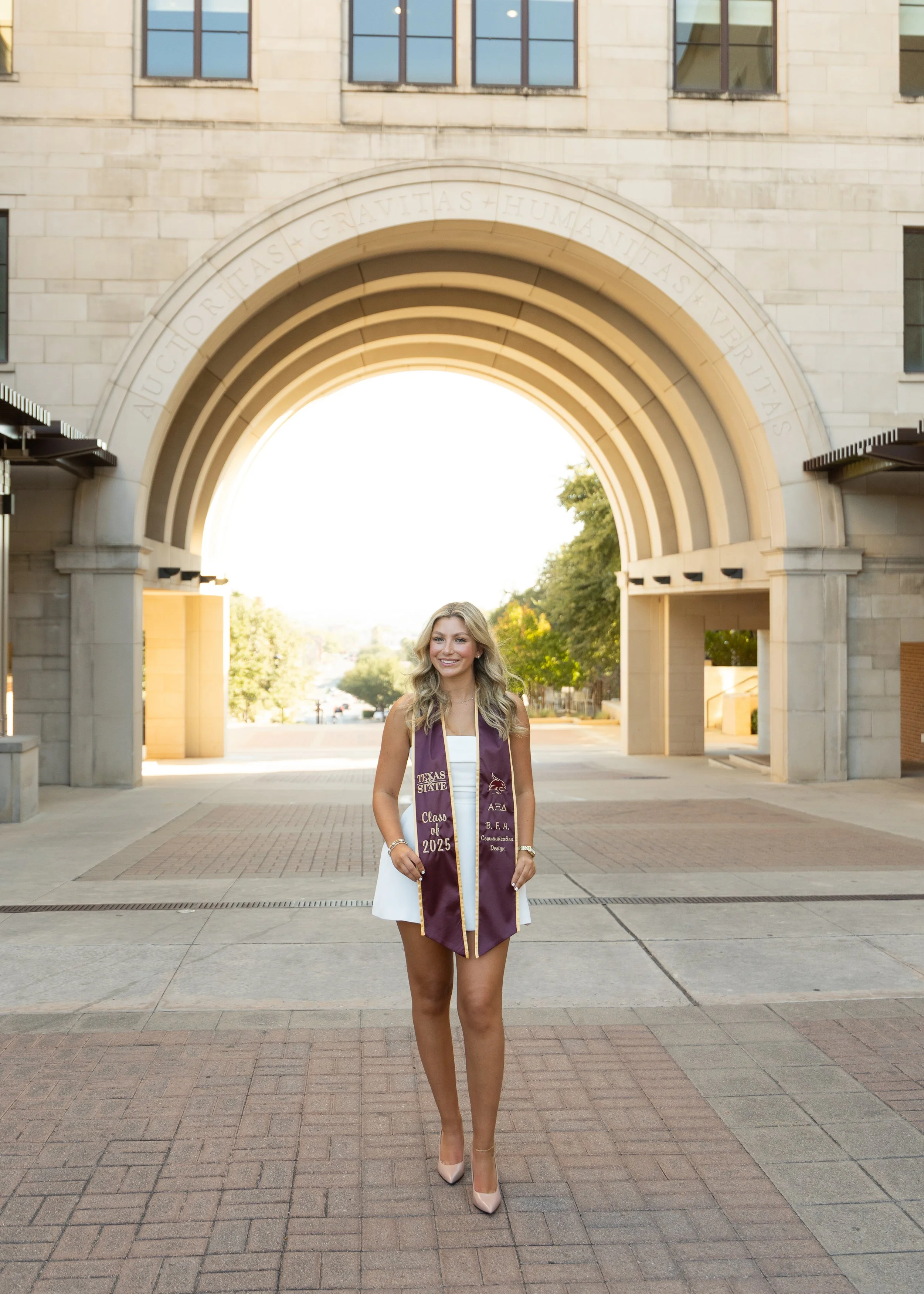A young woman in a white dress and beige high heels standing in front of an archway on a college campus, wearing a graduation sash and stole, smiling at the camera.