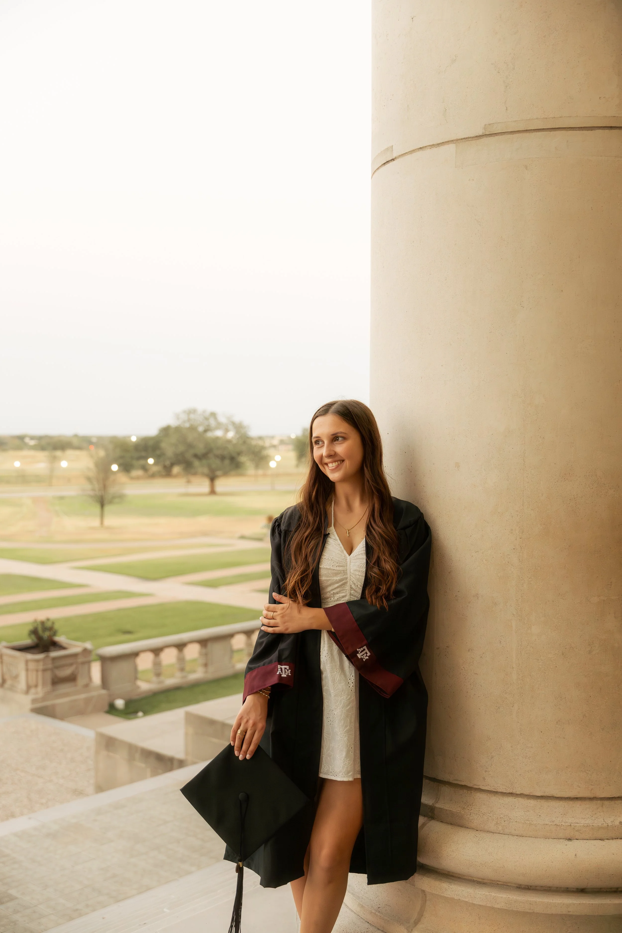 A young woman in a white dress and graduation gown holding a cap, standing against a large stone column with a campus outdoor scene in the background.