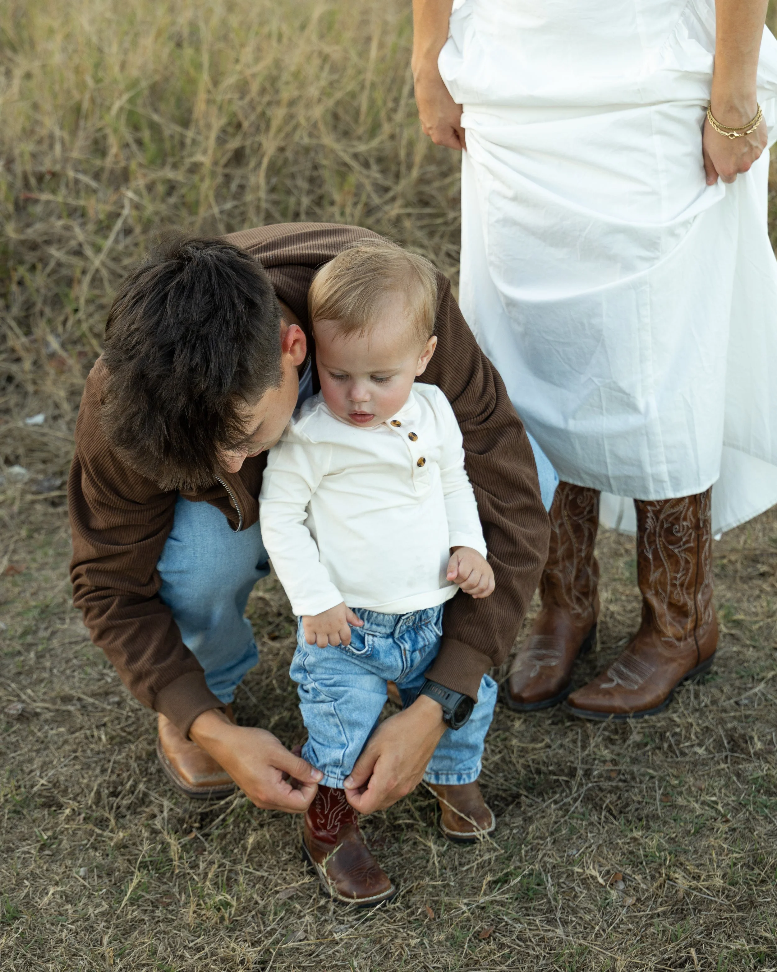A man helping a young boy put on brown cowboy boots outdoors, while a woman standing nearby adjusts her own boots.